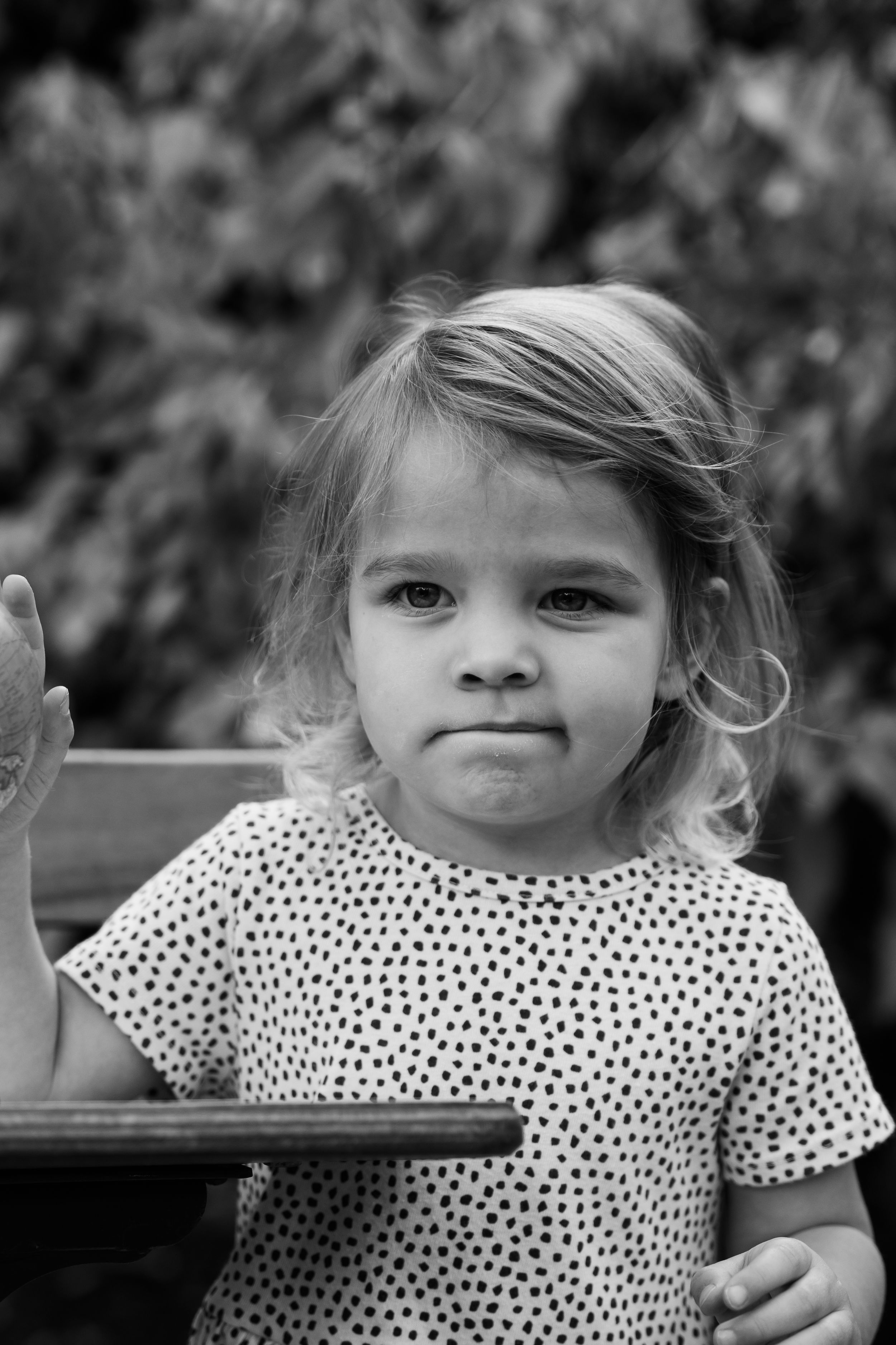 Black and white portrait of a young toddler girl in a polka dot dress, sitting outdoors with a serious expression.