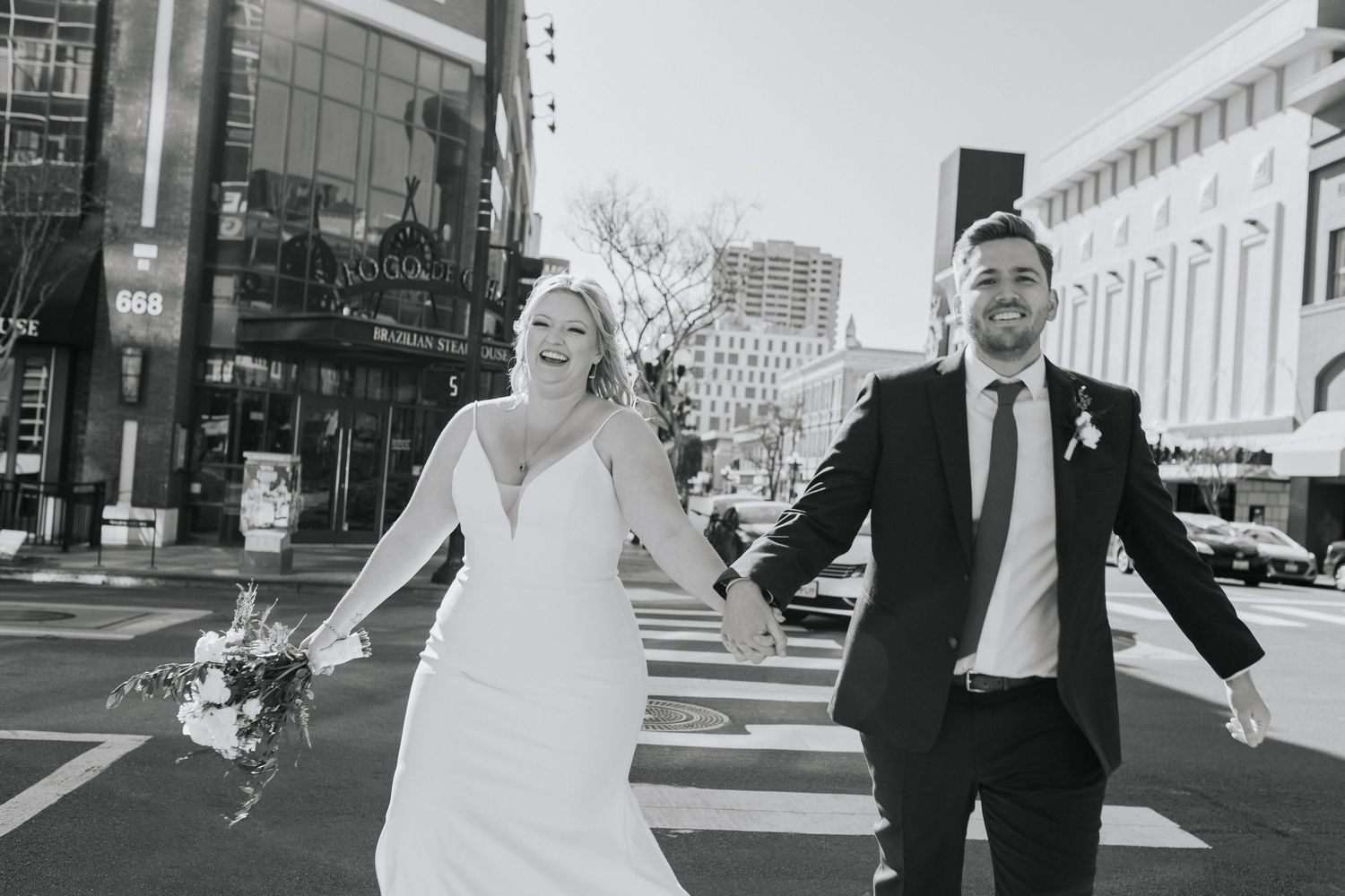 A man and woman holding hands, wearing wedding attire, run across a street in San Diego.