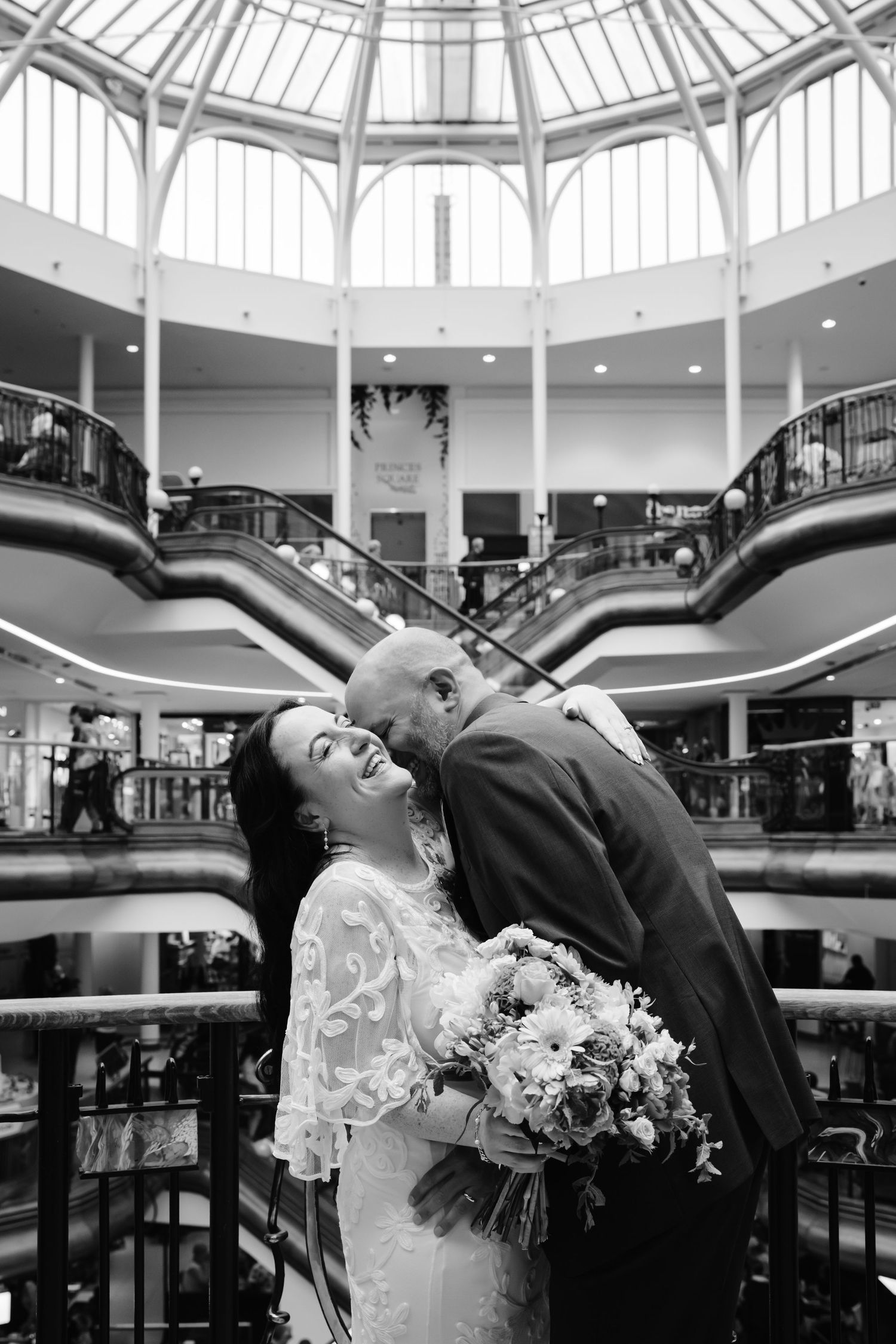 Black and white wedding photo of bride and groom sharing a romantic moment in an elegant mall atrium with escalators.