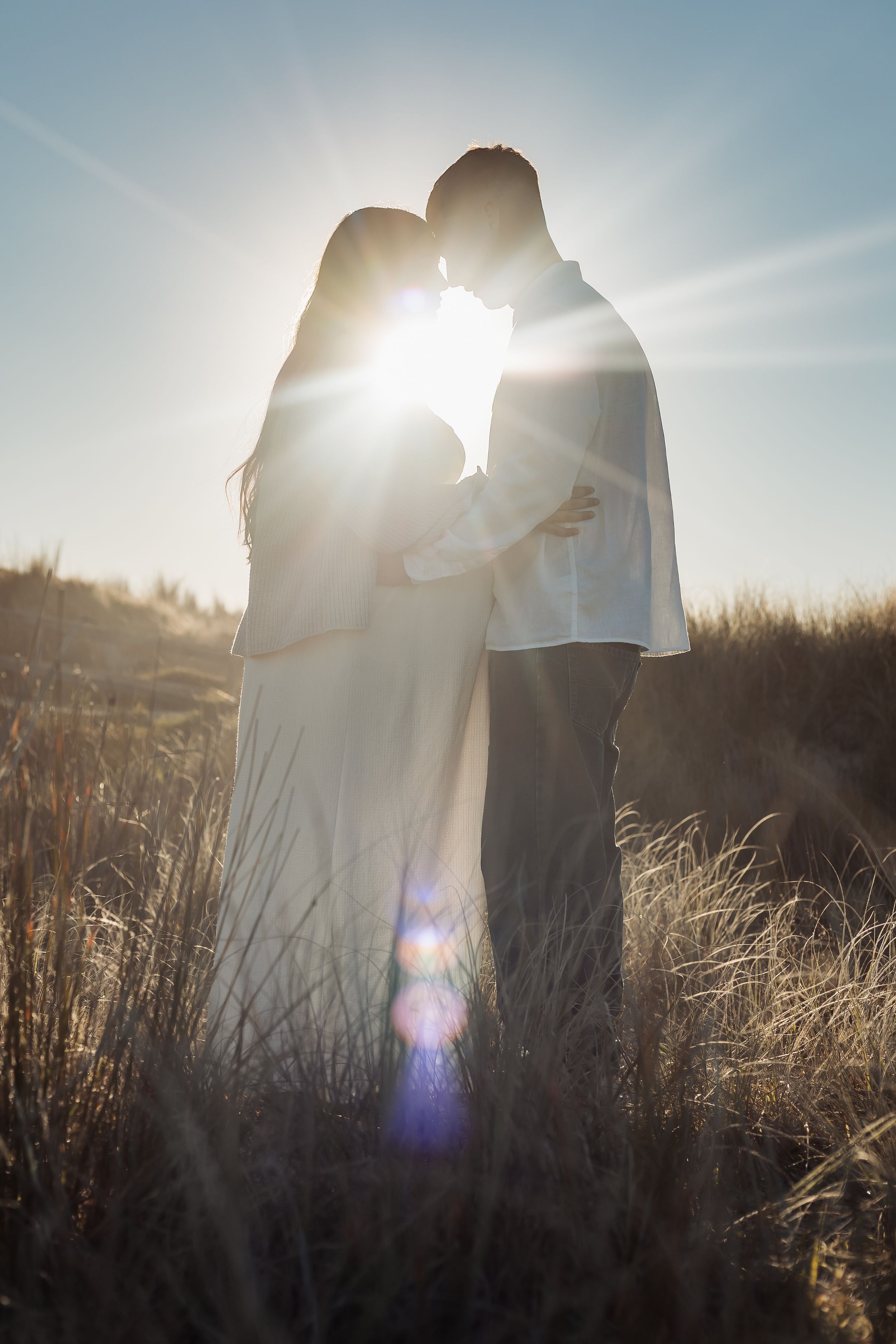 Couple embracing in golden field with sun flare at sunset, silhouetted against blue sky.