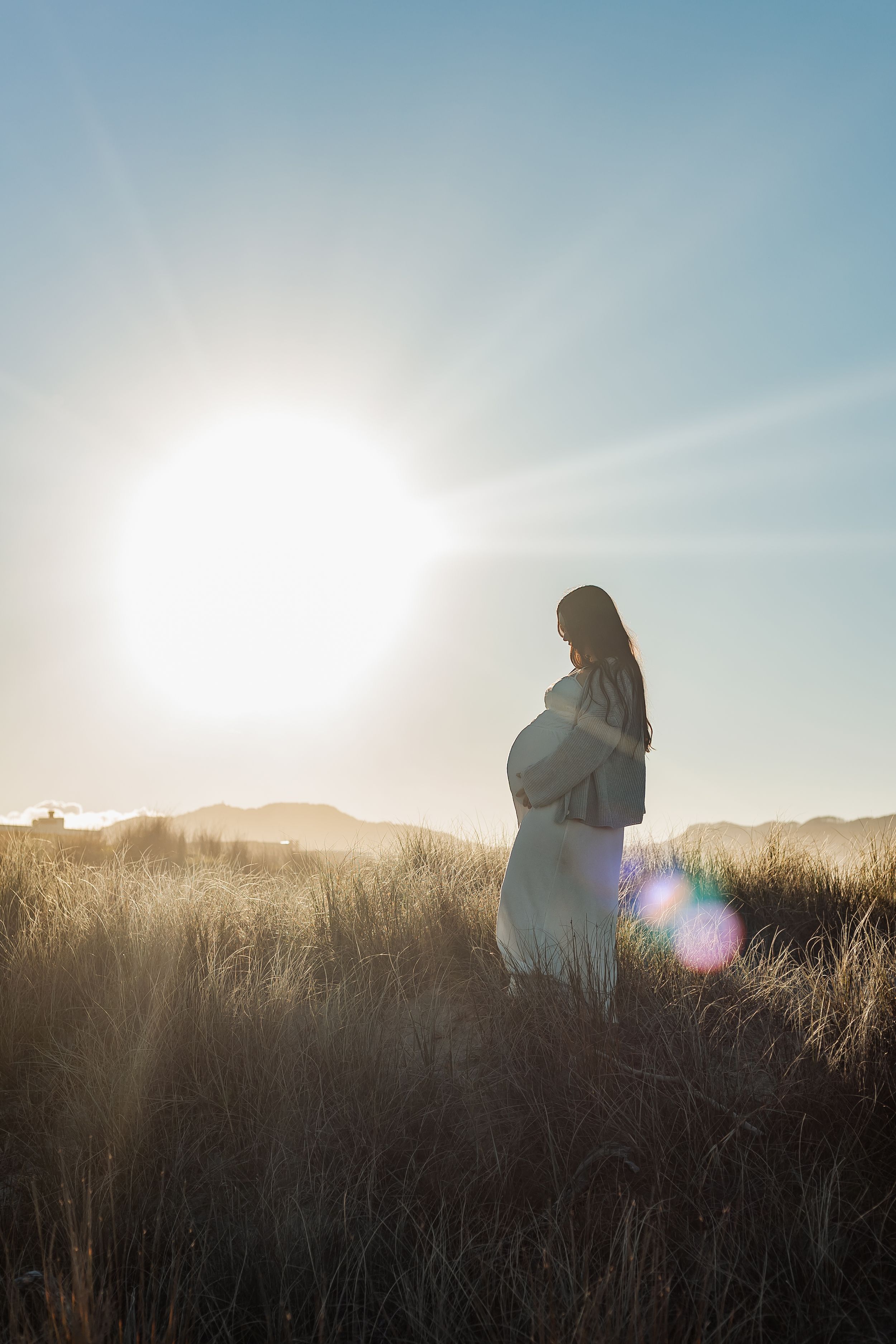 Pregnant woman standing in golden field at sunset, cradling her baby bump with mountains in the background.