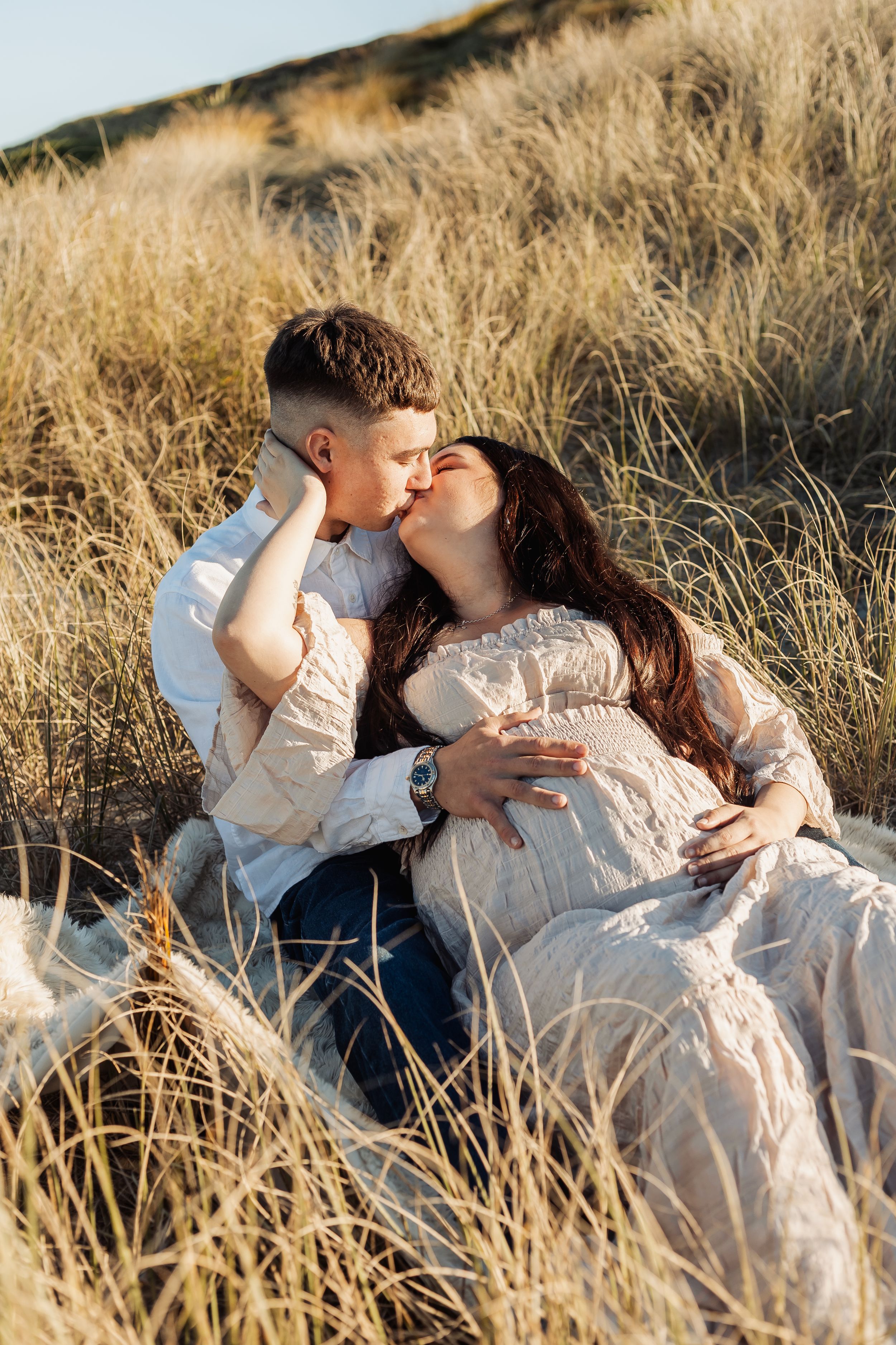 Couple kissing in golden grass field, pregnant woman in lace dress, romantic maternity photo shoot outdoors.