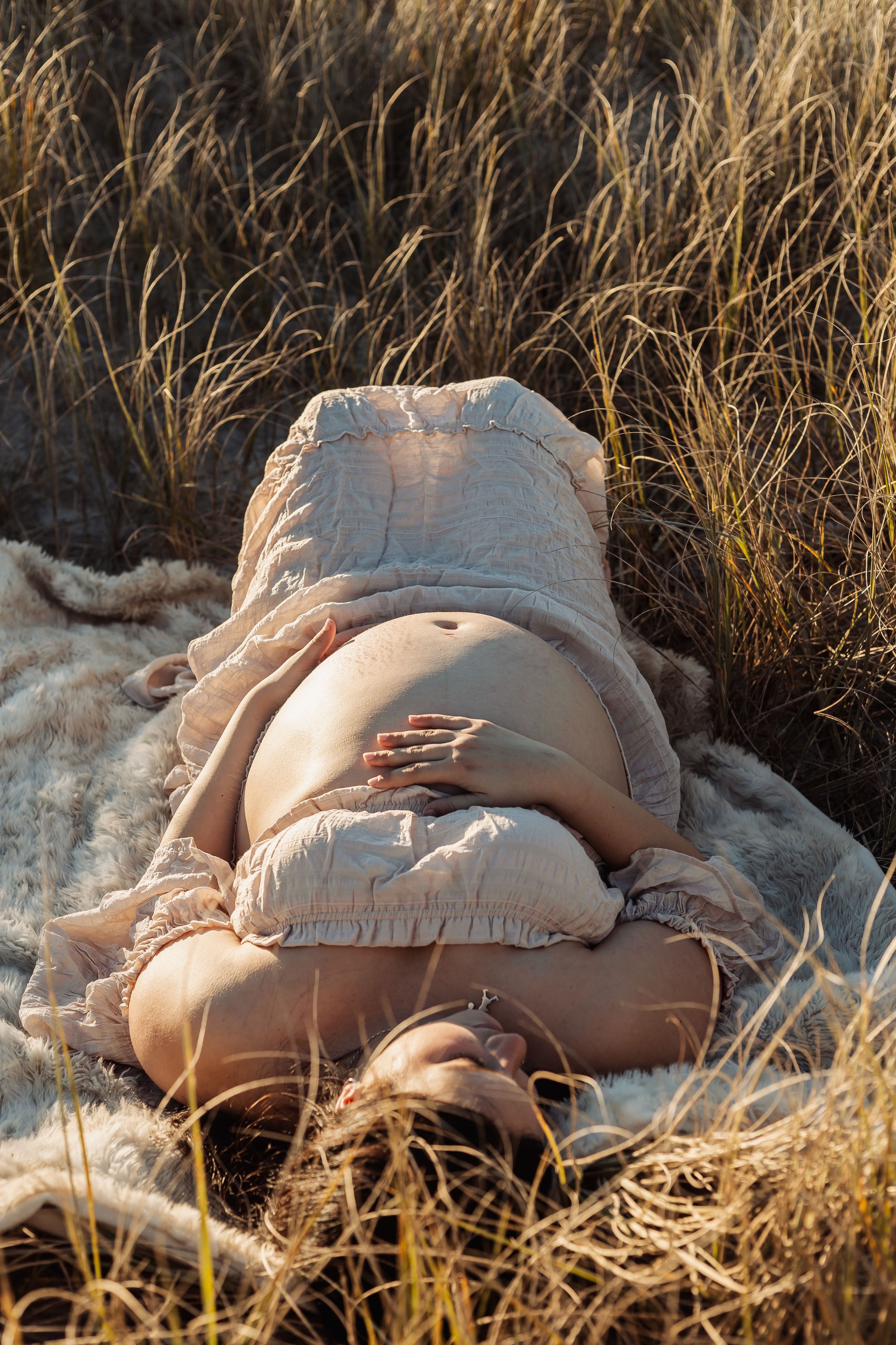 Pregnant woman lying on a blanket in golden grass, cradling her bare baby bump in warm sunlight.