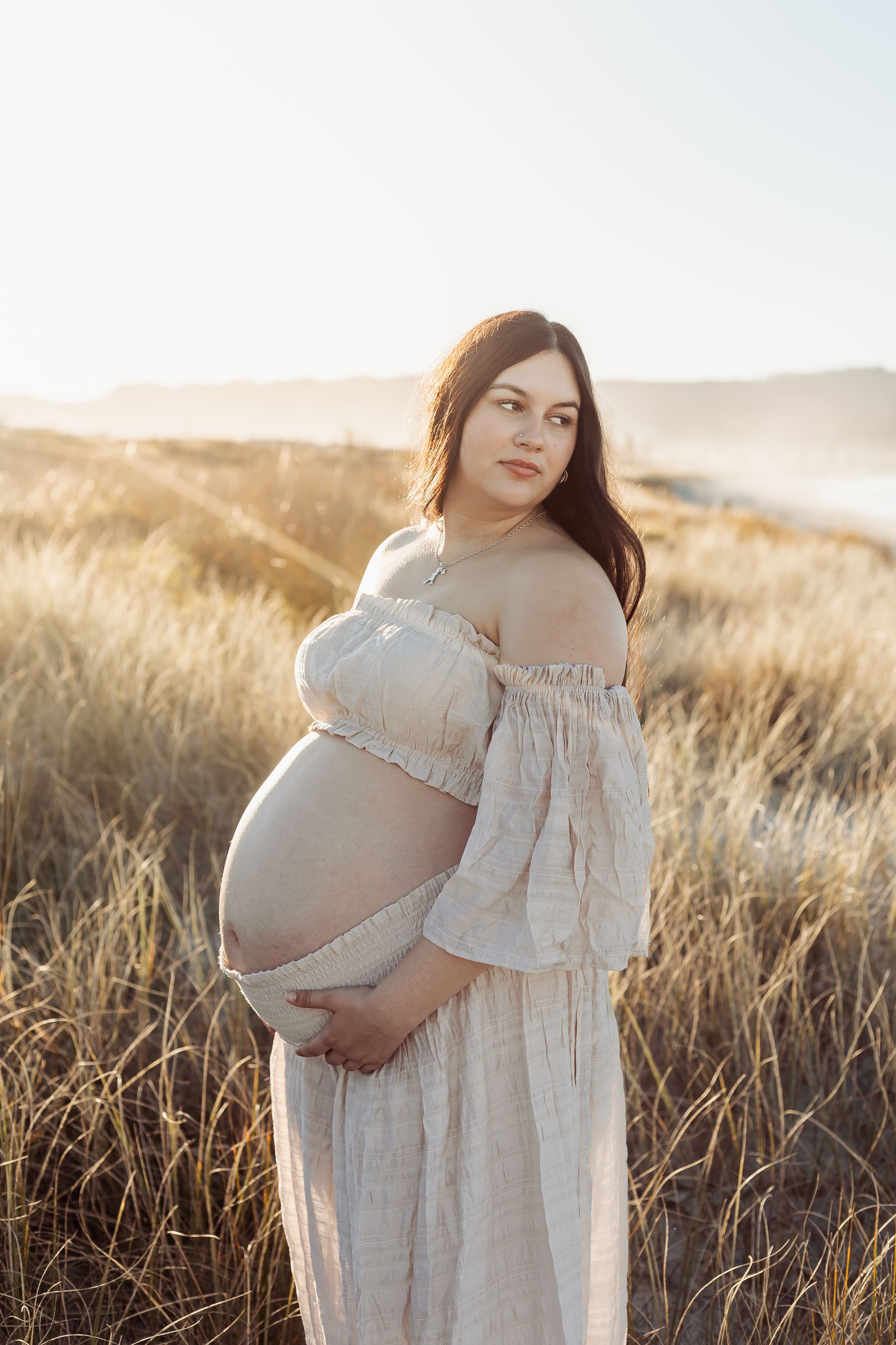 Pregnant woman in off-shoulder beige dress holding belly, standing in golden grass field at sunset.
