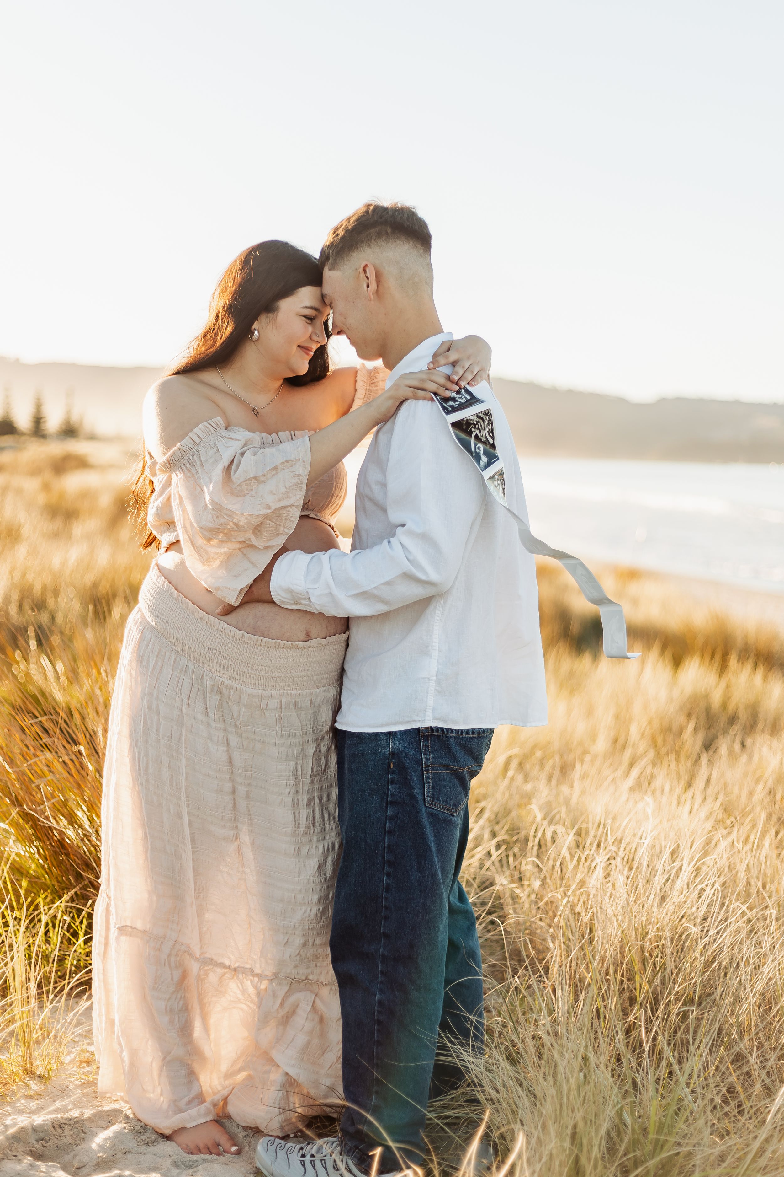 Pregnant couple embracing in golden beach dunes at sunset, woman in blush dress, man holding ultrasound photo.