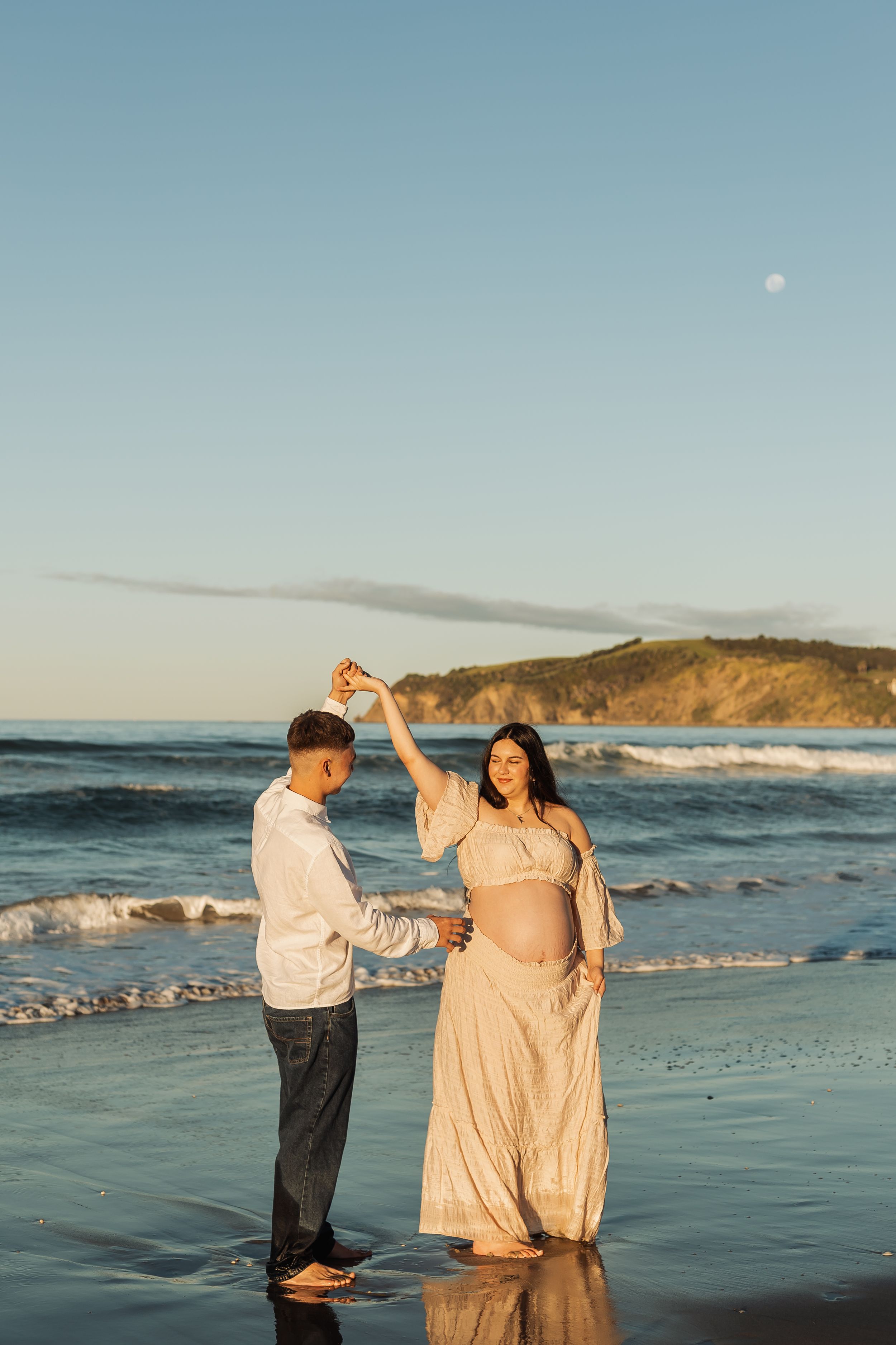 Couple dancing barefoot on a beach at sunset, pregnant woman in flowing cream dress, waves and cliffs in background.