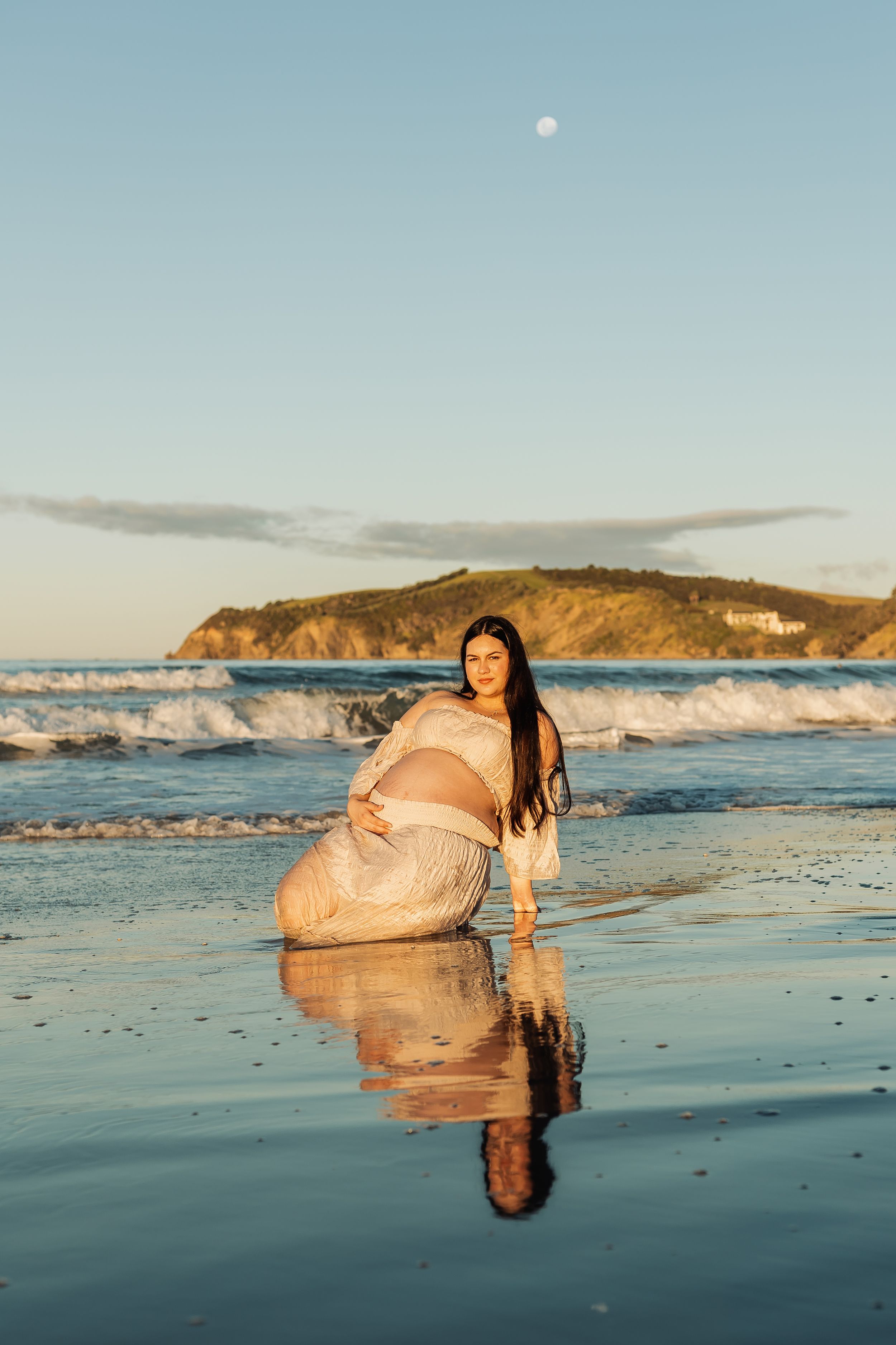 Pregnant woman in white dress kneeling on reflective beach at golden hour with ocean waves and moon.