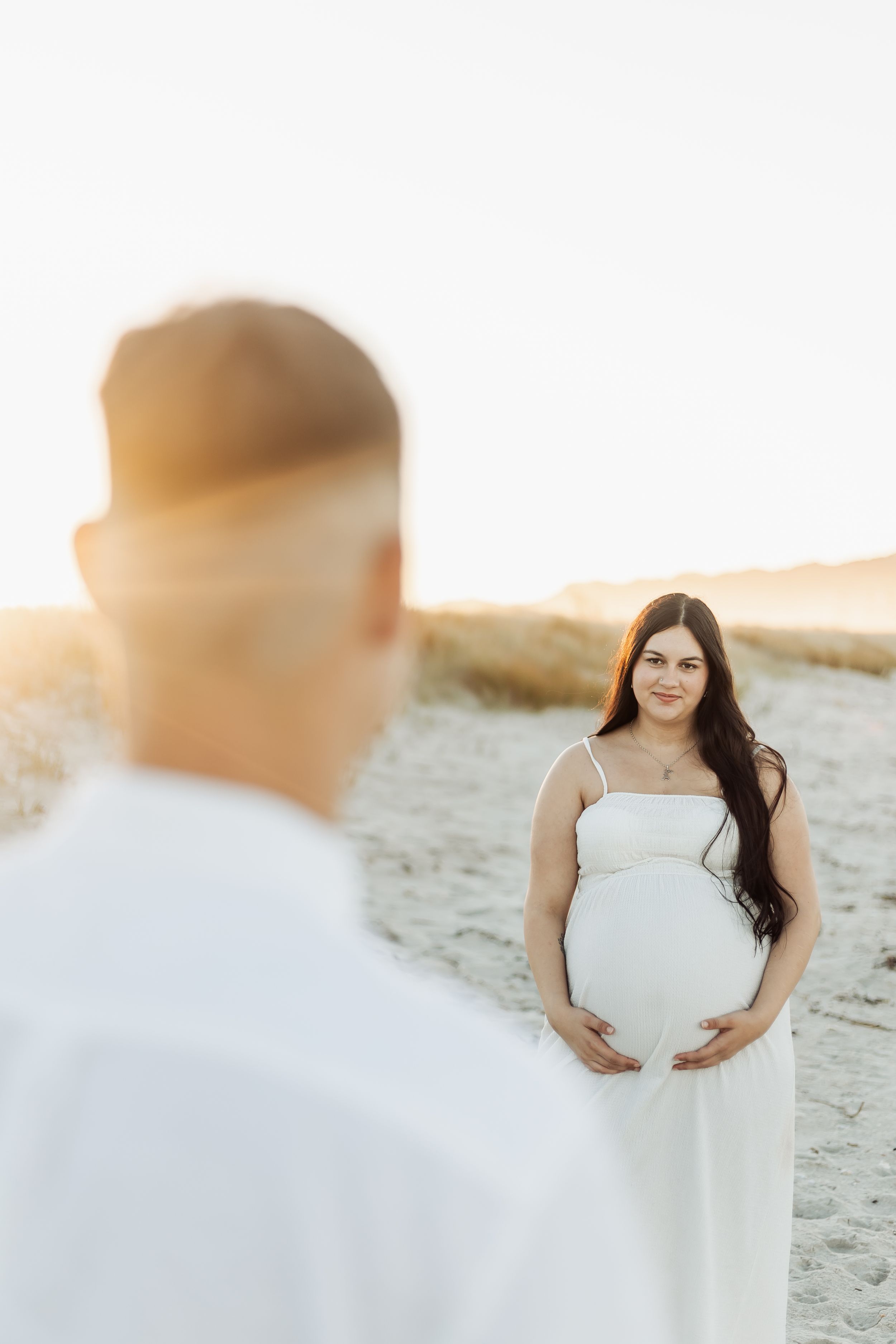 Pregnant woman in white dress holds belly on sandy beach, man in white shirt stands in foreground at golden hour.