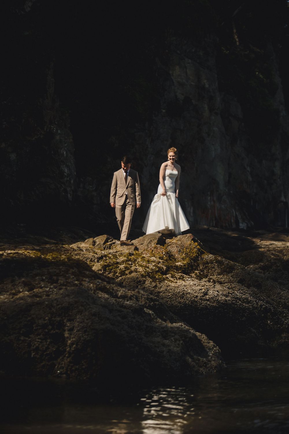 Bride in white gown and groom in tan suit stand on rocky shore with dark cave backdrop.