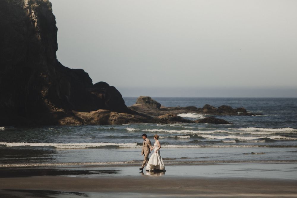 A couple in wedding attire walks along a scenic beach with dramatic rock formations and ocean waves in the background.