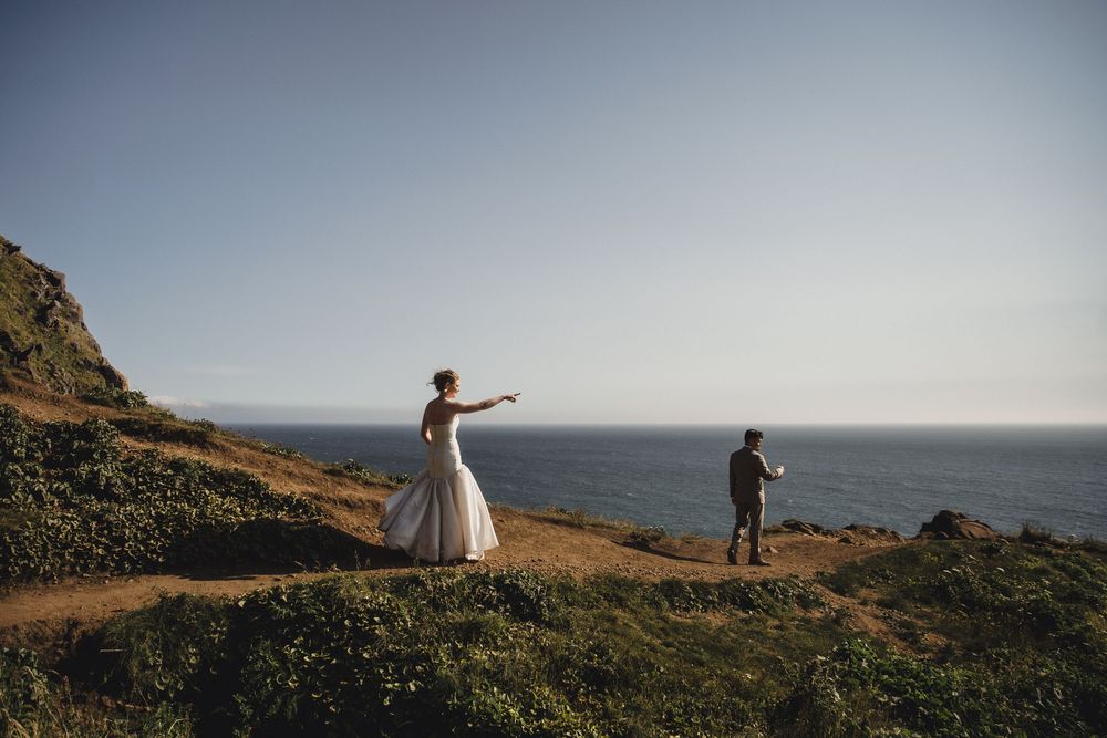 Bride in white gown points toward ocean while groom stands nearby on scenic coastal cliffside wedding photo.