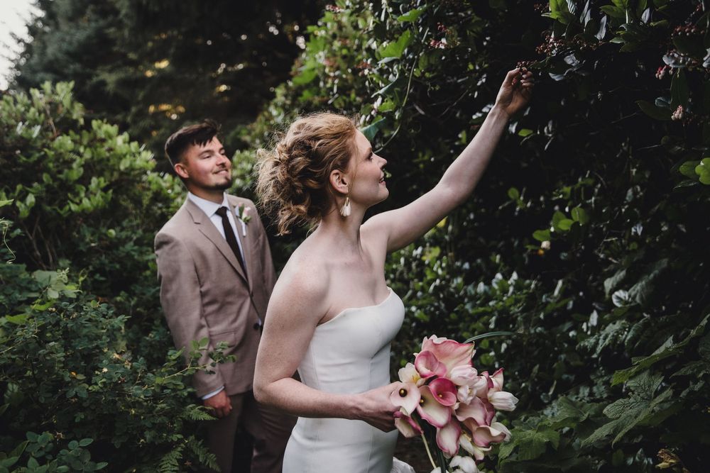 Bride holding pink bouquet reaches toward greenery while groom in tan suit smiles beside her outdoors.
