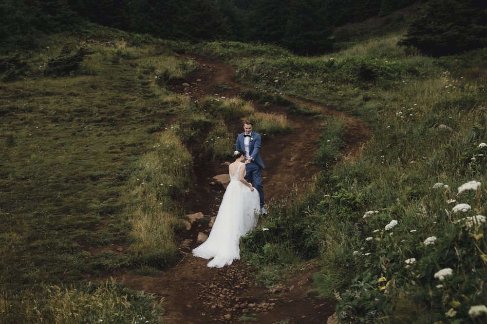 Bride and groom stand on a scenic mountain trail surrounded by lush green hills on their wedding day.