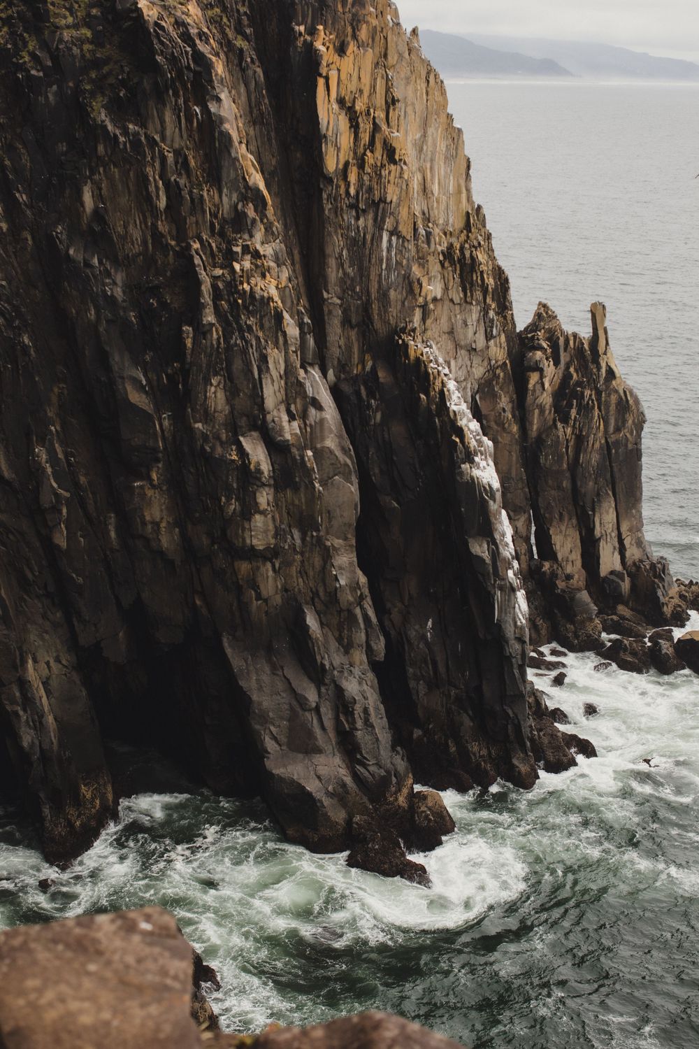 Dramatic rocky coastal cliffs with jagged stone formations rising from turbulent ocean waves below on an overcast day.