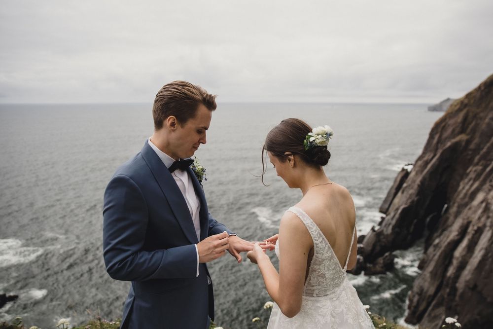 Groom placing wedding ring on bride's finger at dramatic coastal cliffs with ocean backdrop on overcast day.