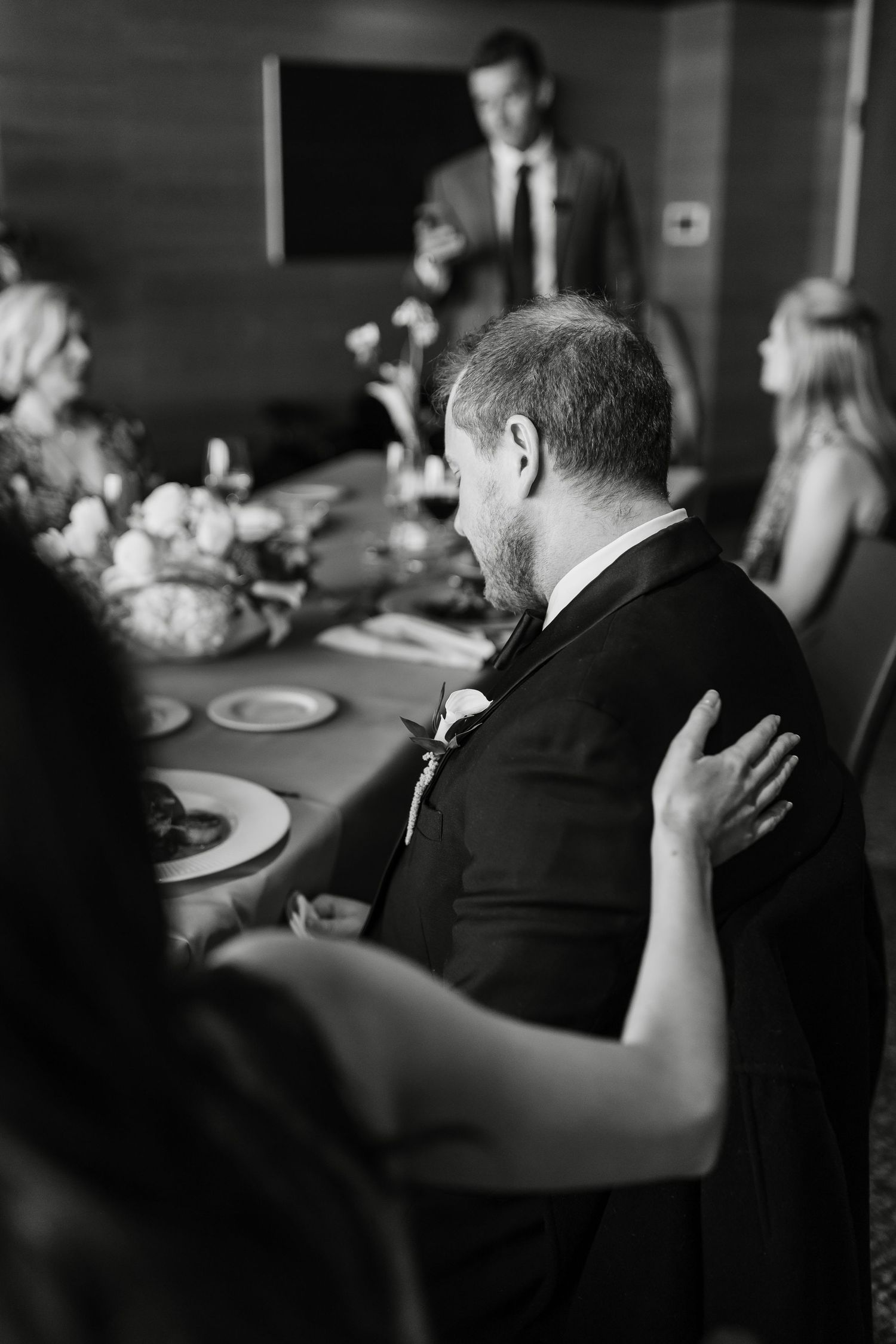 Black and white wedding reception photo of groom listening to a toast at an elegant dinner table.