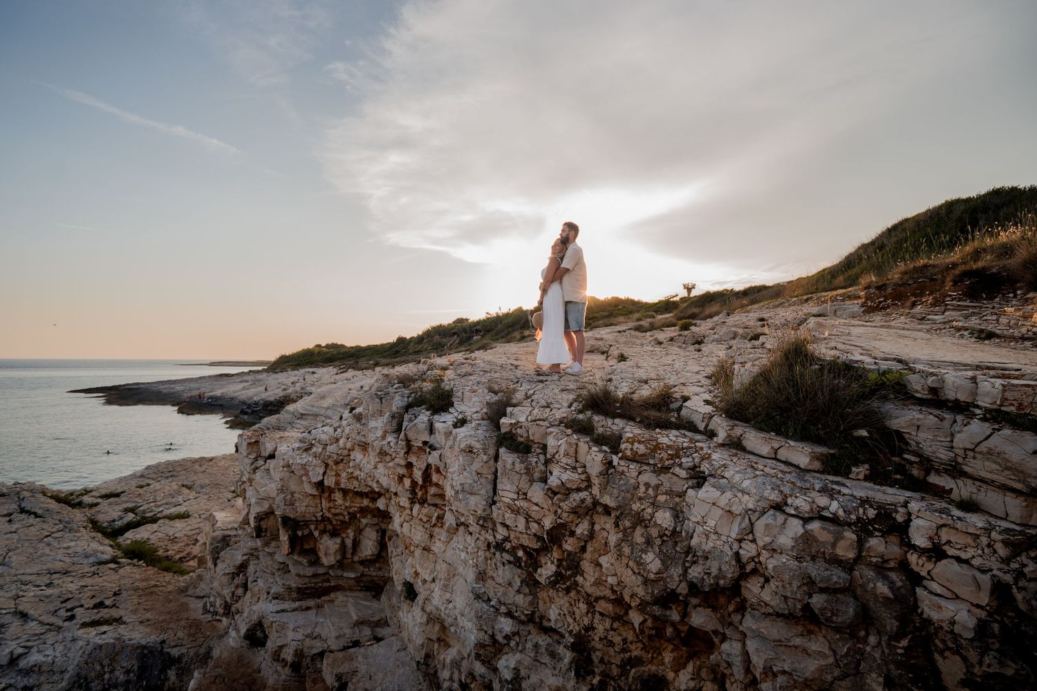 Woman in white dress standing on rocky coastal cliffs at sunset with calm sea and green hills behind her.