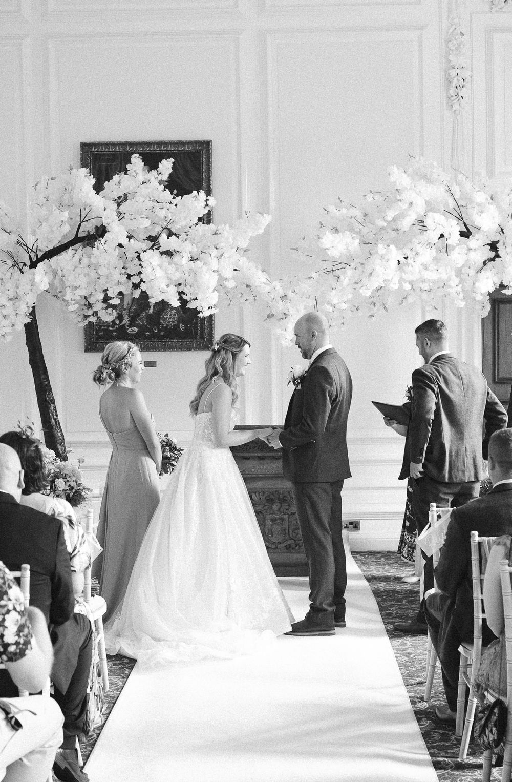 Black and white photo of a bride and groom exchanging vows at an elegant wedding ceremony with white cherry blossom trees.