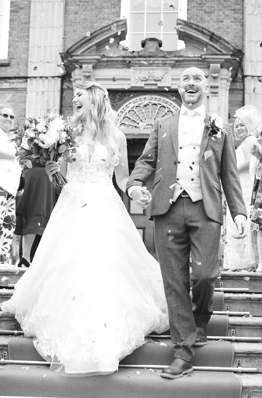 Joyful bride and groom laughing on church steps during confetti shower in black and white wedding photo.