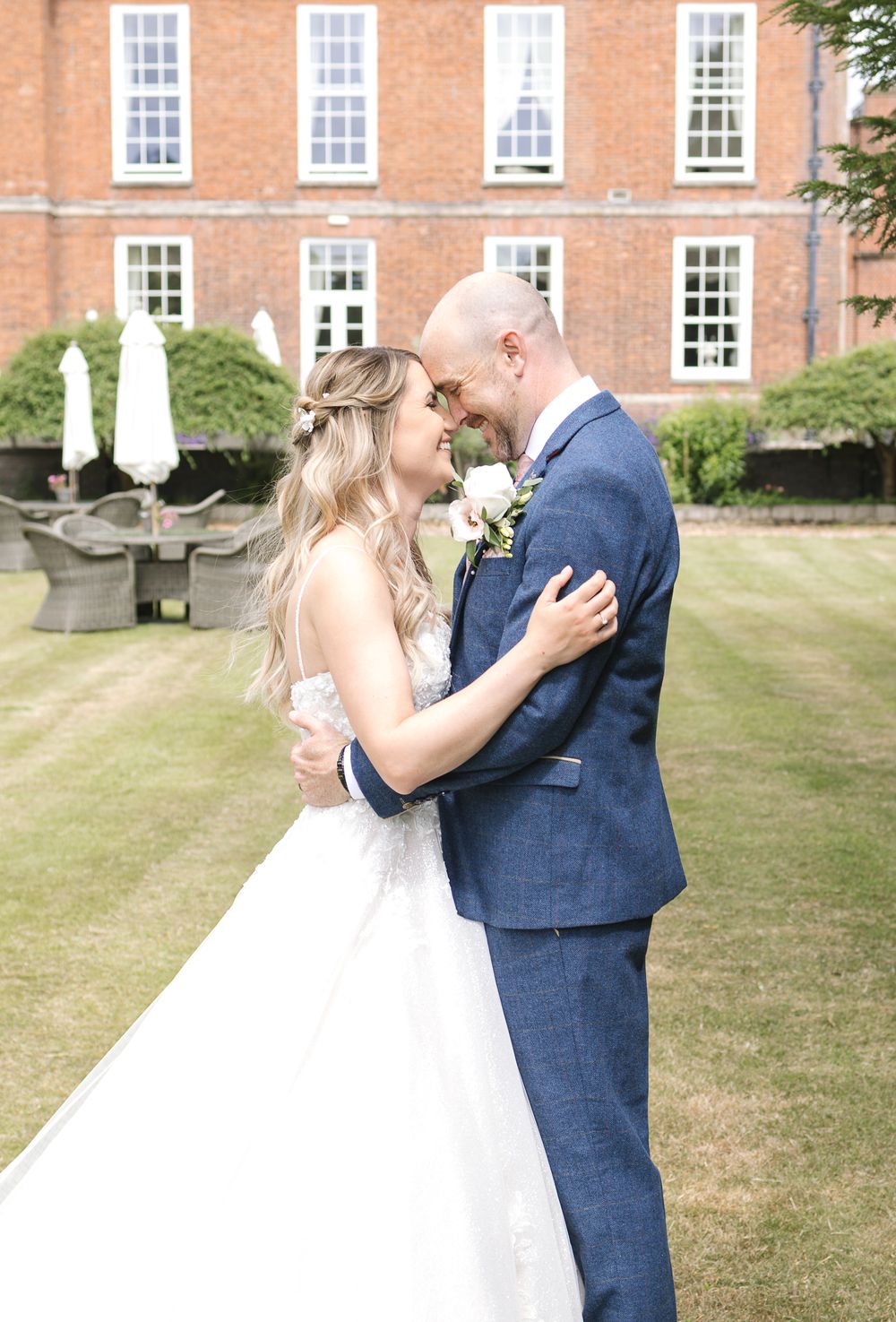 Bride and groom sharing romantic moment in sunny garden outside brick wedding venue.
