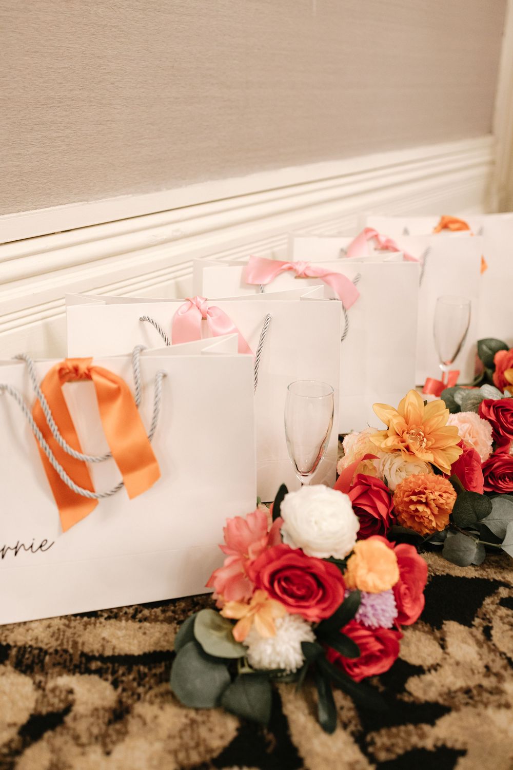 White gift bags with pink and orange ribbons lined up with colorful floral bouquets and champagne flutes.
