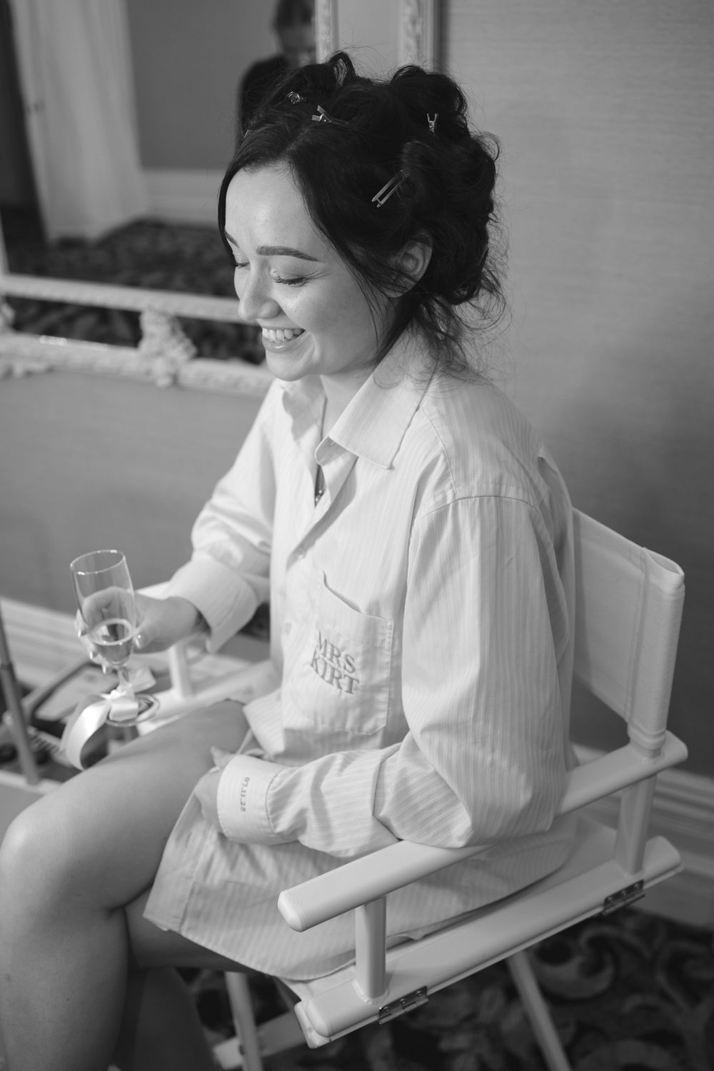 Black and white photo of a smiling bride in a monogrammed robe holding champagne while seated in a white chair.