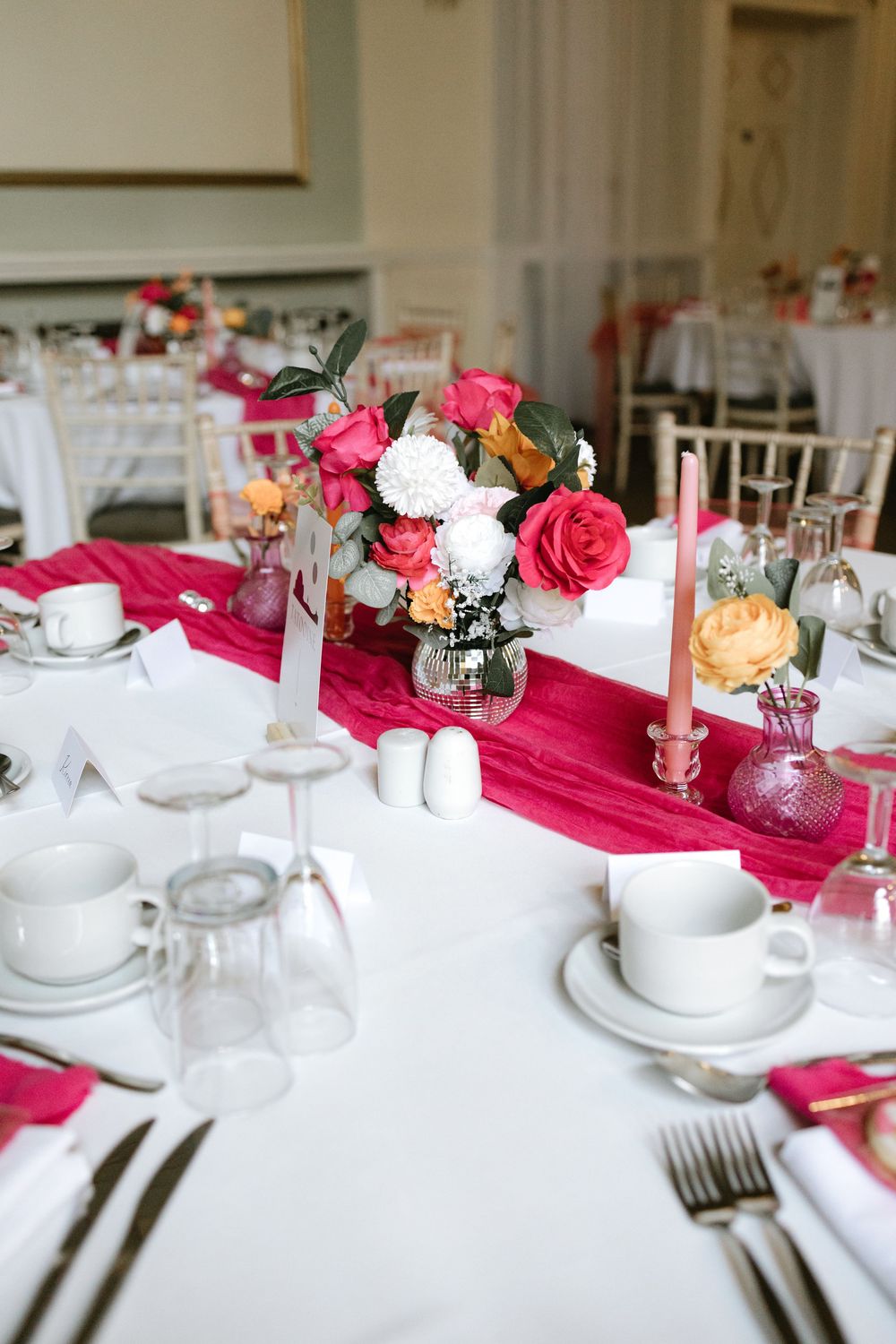 Elegant wedding reception table with hot pink runner, floral centerpiece of roses, white dinnerware, candles, and crystal glassware.