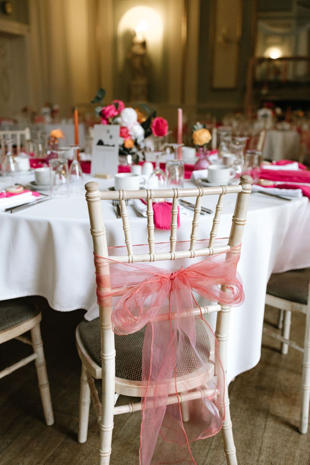 Elegant wedding reception chair with pink organza bow, set at a white tablecloth table with floral centerpieces.