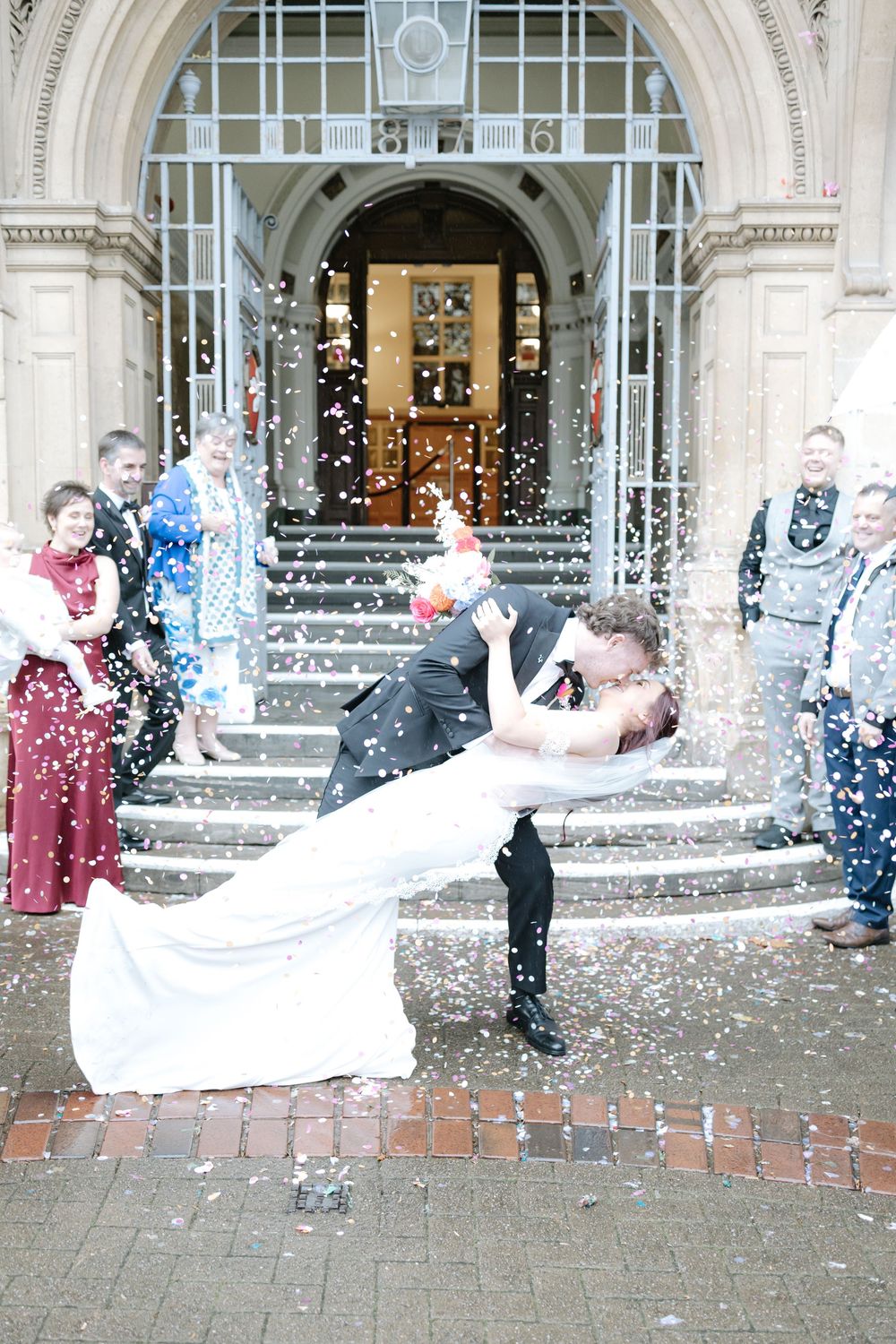 Bride and groom share romantic dip kiss outside grand venue entrance with confetti shower and wedding guests celebrating.