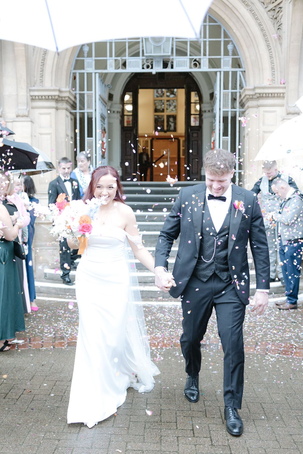 Newlywed couple walking out of wedding venue as guests throw confetti, bride in white gown holding bouquet.