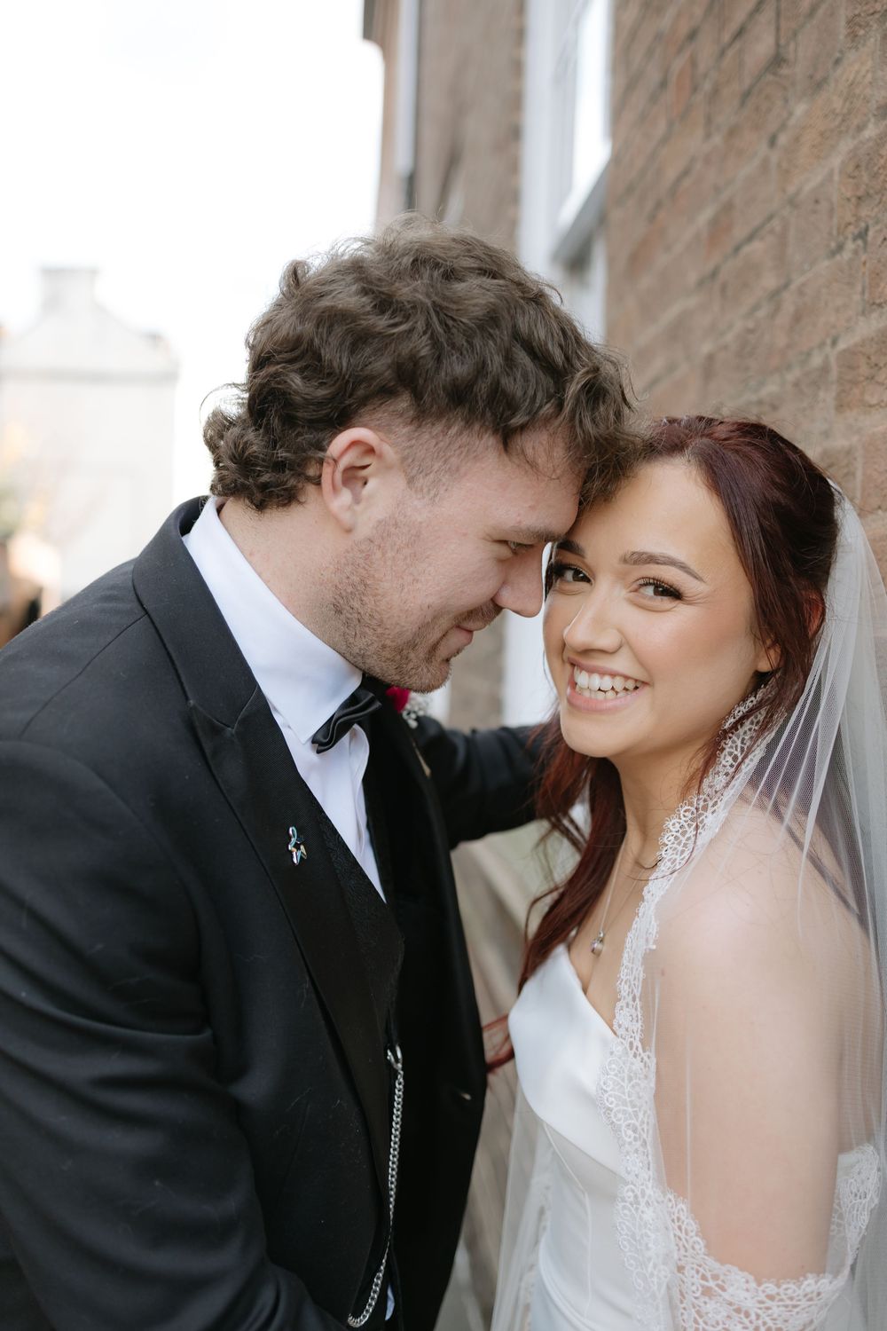 A smiling bride in a white veil leans against a brick wall as her groom in a black suit tenderly touches foreheads with her.