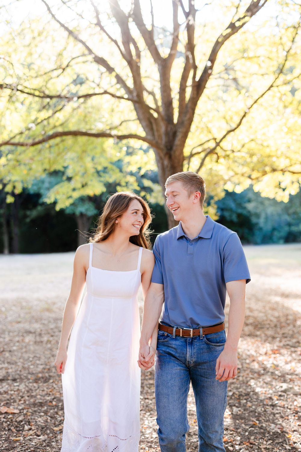 A smiling couple holds hands while walking in a sunlit park, with a large tree and golden foliage in the background.