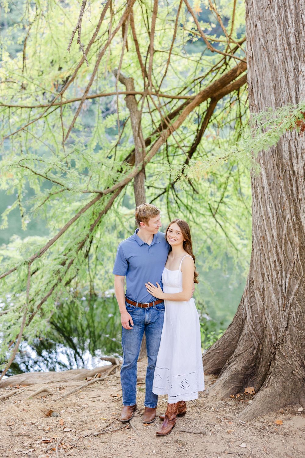 Couple embracing under a large tree outdoors, man in blue shirt and jeans, woman in white sundress, lush green background.