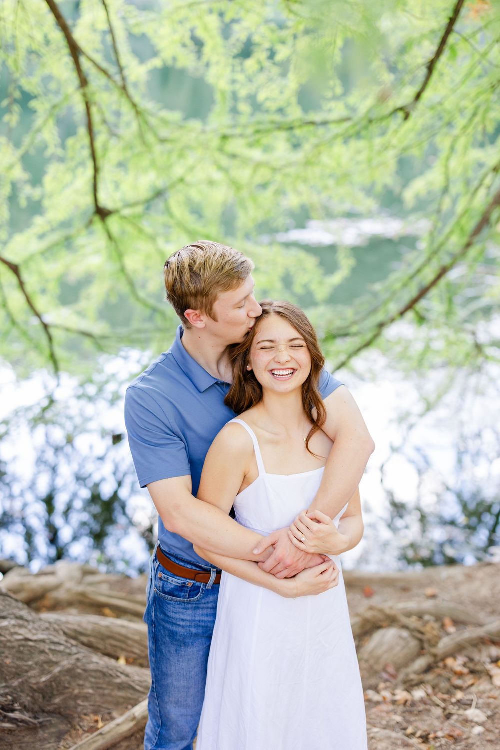 Couple embracing and laughing under green trees by a lake, man kissing woman's cheek, she wears white dress.