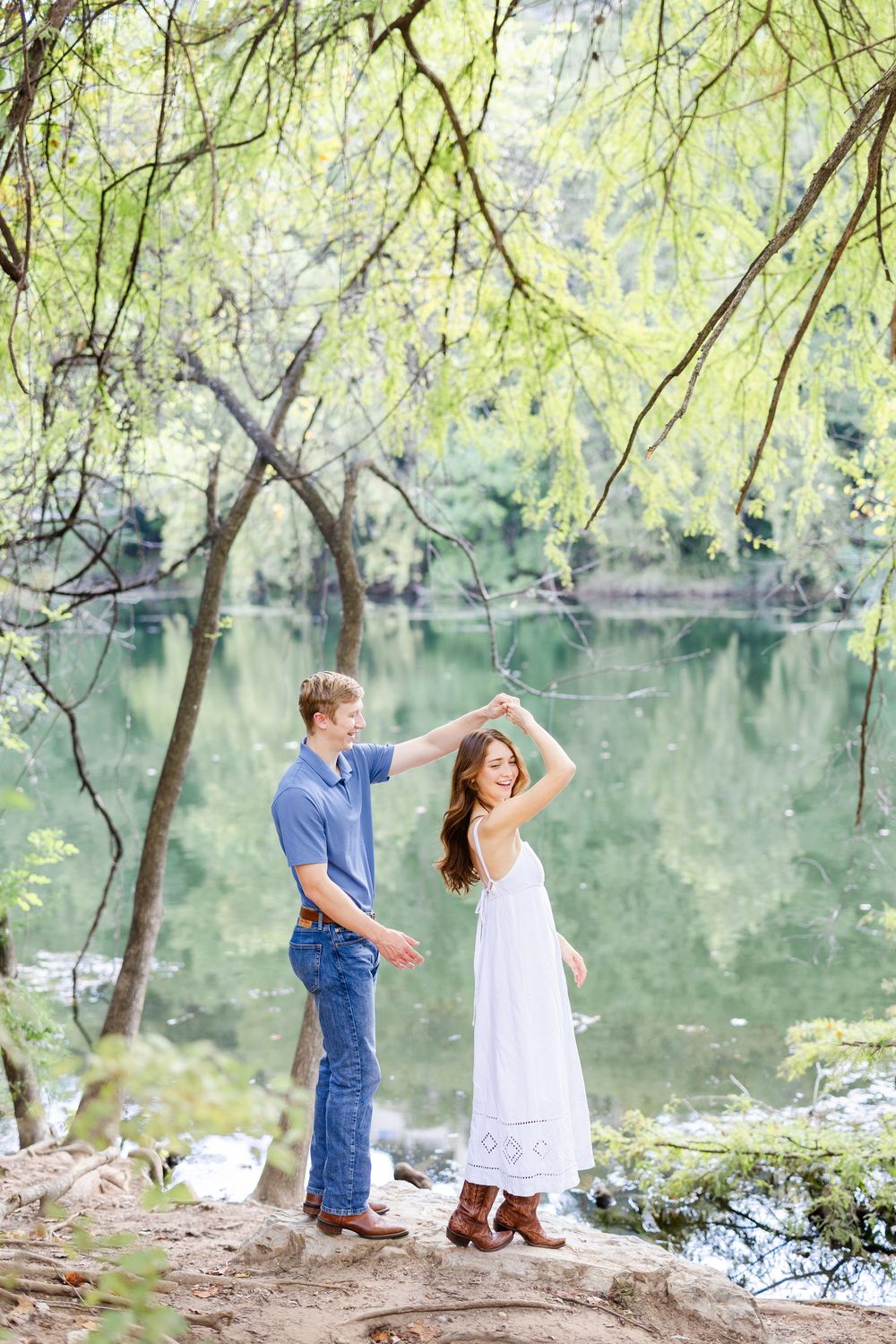 Couple dancing outdoors near a serene green lake, man in blue shirt twirling woman in white dress under weeping willow trees.