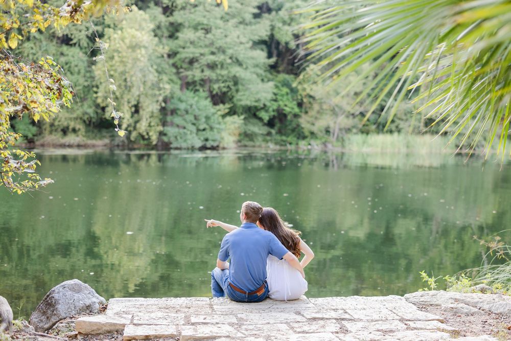 Romantic couple sitting together on a stone ledge pointing at a peaceful green lake outdoors.