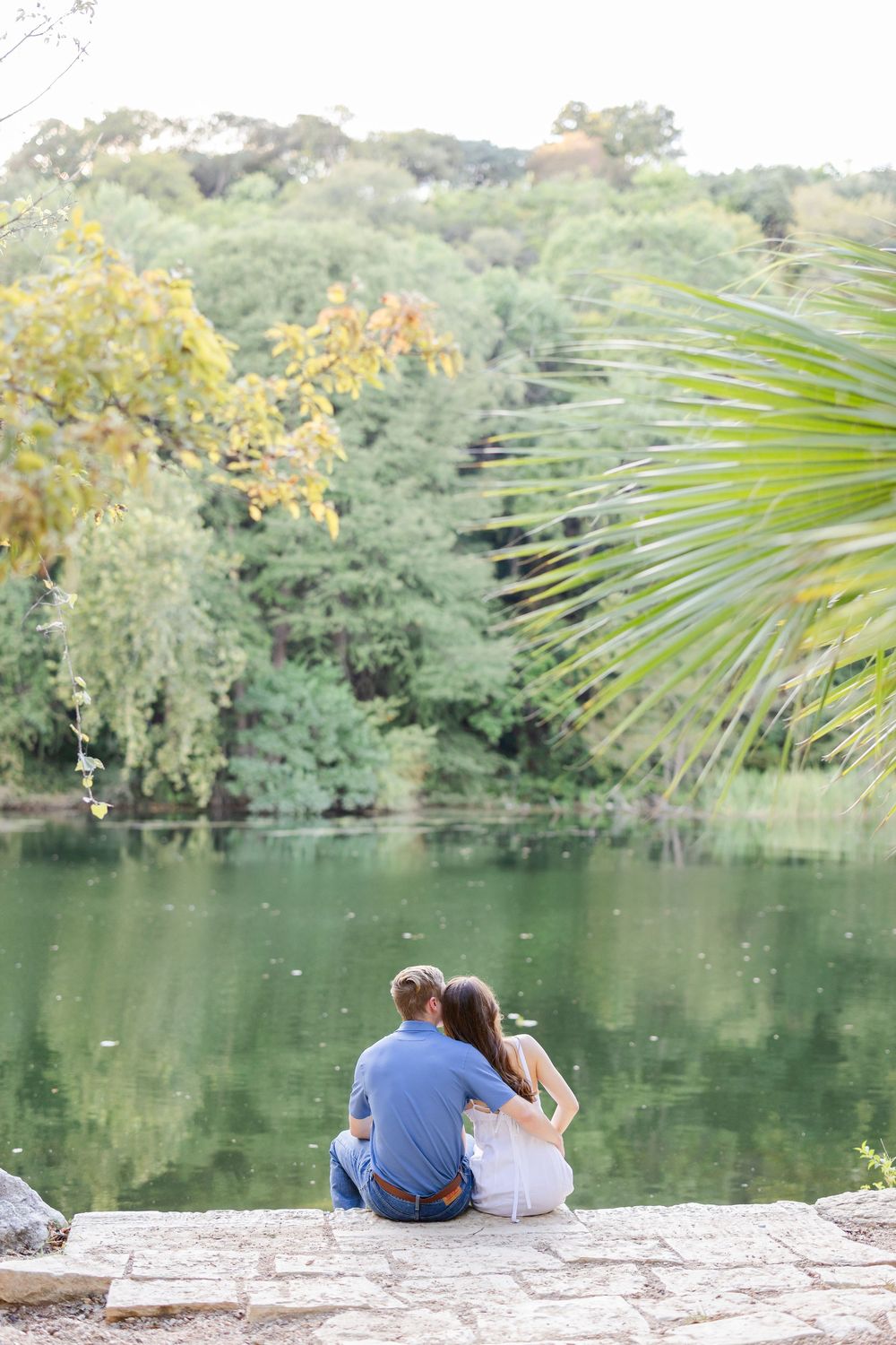 Couple sitting together on stone ledge overlooking a serene green lake surrounded by lush trees and a palm frond.