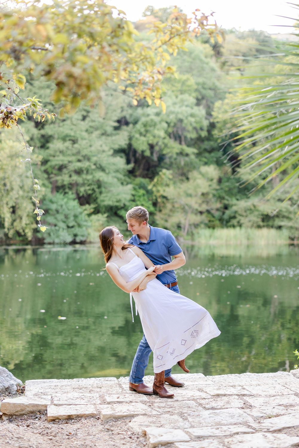 Happy couple dancing and laughing together on stone steps beside a tranquil green lake outdoors.