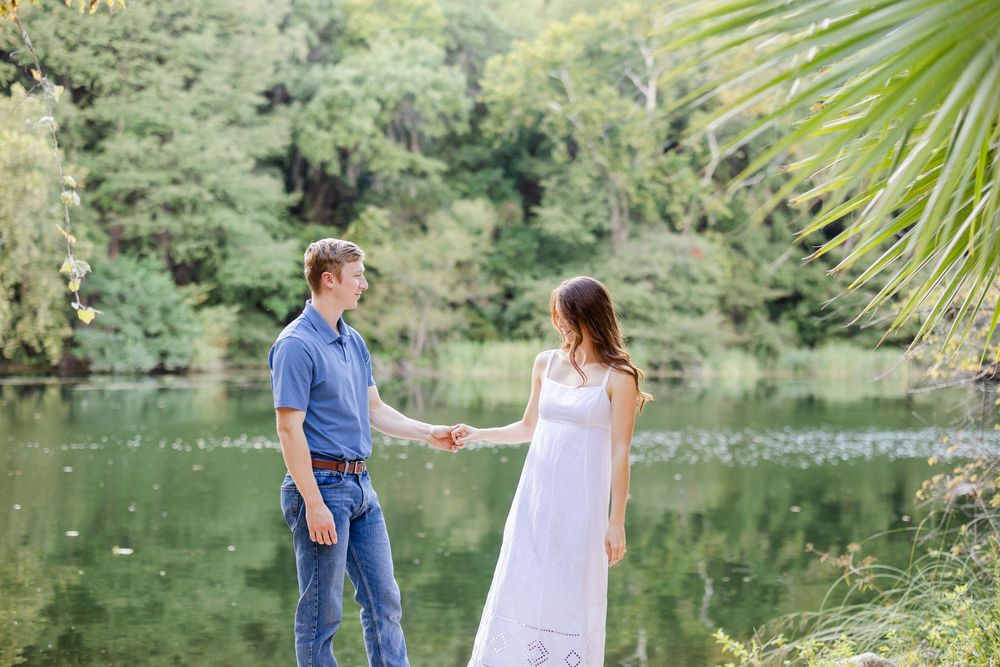 Couple holding hands near a serene lake surrounded by lush green trees, woman in white dress, man in blue jeans.