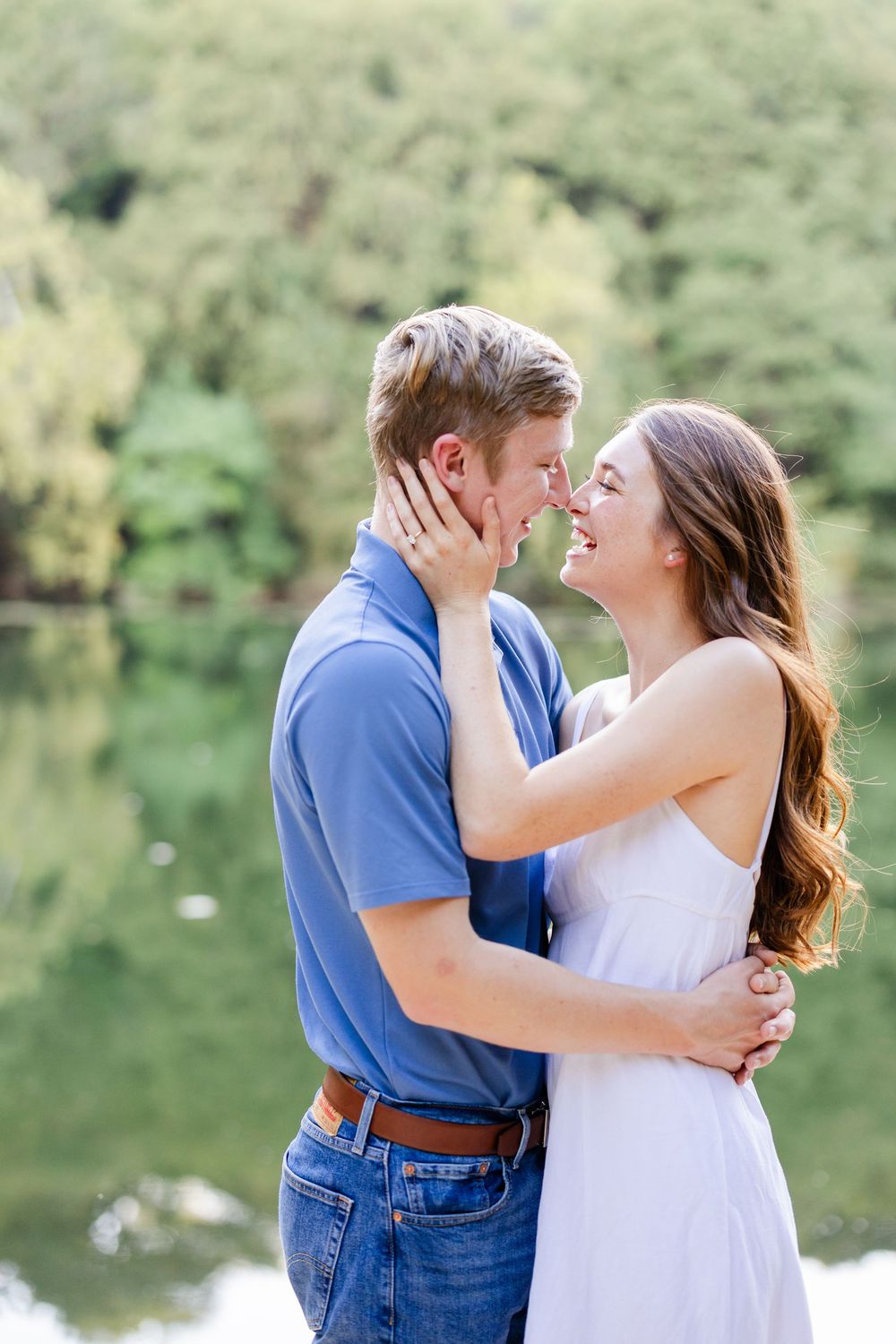 A happy couple shares a romantic moment by a lake, the woman in a white dress holding the man's face as they lean in for a kiss.