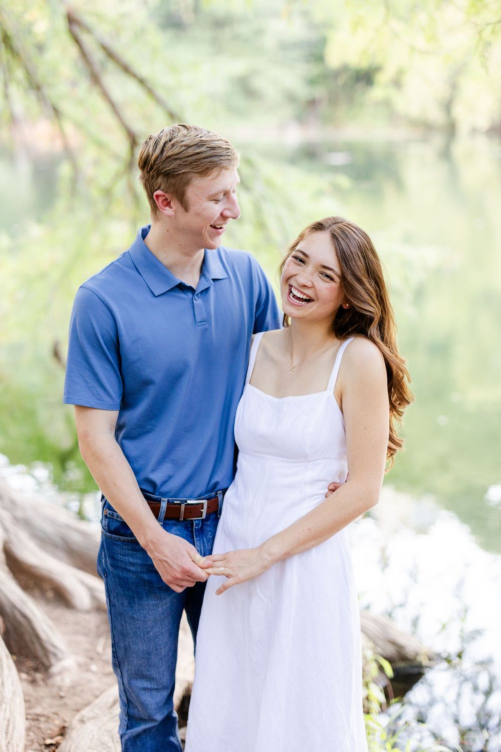 Happy couple laughing together outdoors near a pond, man in blue polo and jeans, woman in white sundress.