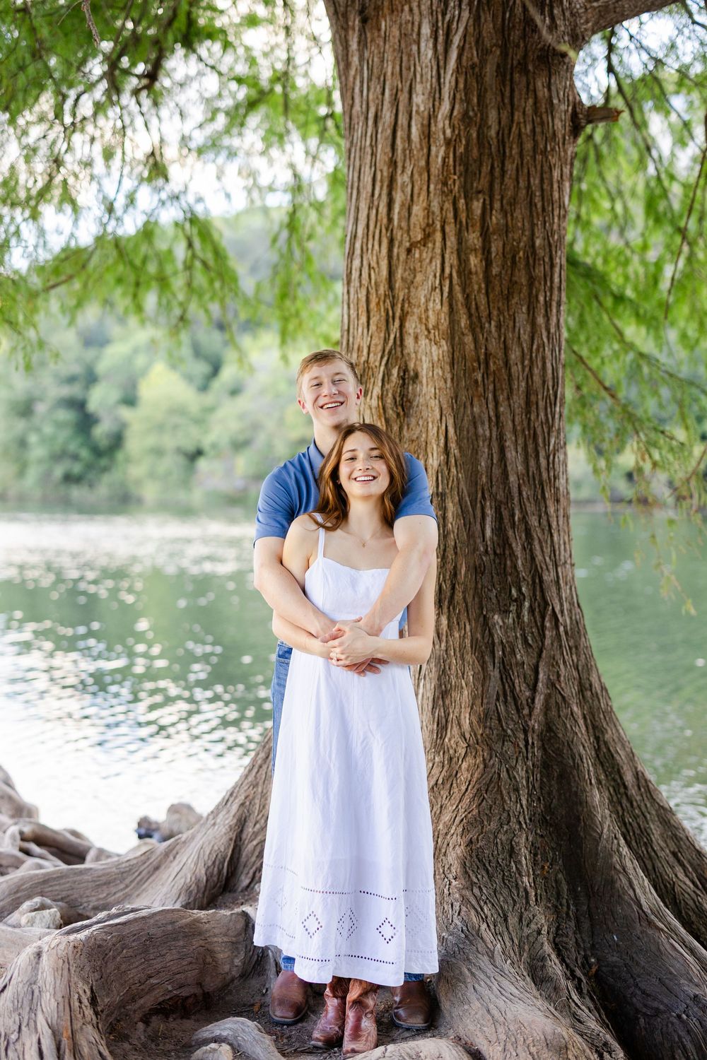 Happy couple embracing by a large tree near a lake, woman in white dress, man in blue shirt, outdoor nature setting.