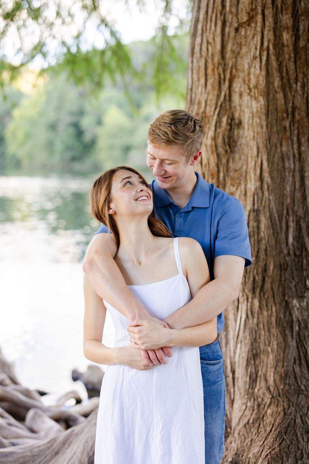 Couple embracing by a tree near a lake, woman in white dress smiling up at man in blue polo shirt.