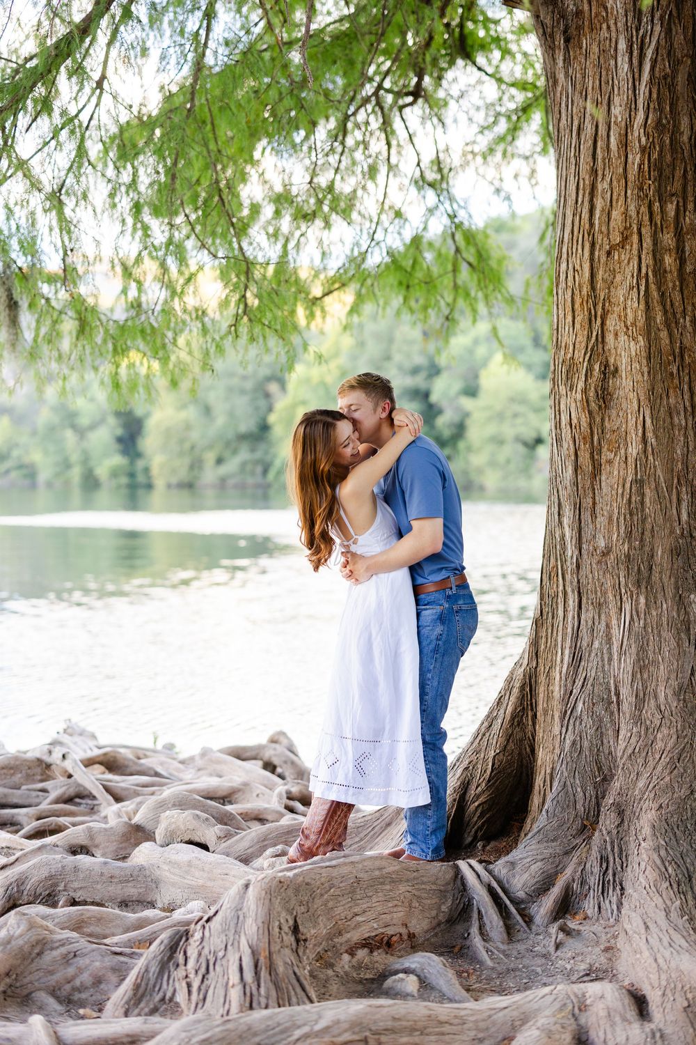 Couple sharing a romantic kiss under a large tree by a serene lake, woman in white dress, man in blue shirt.