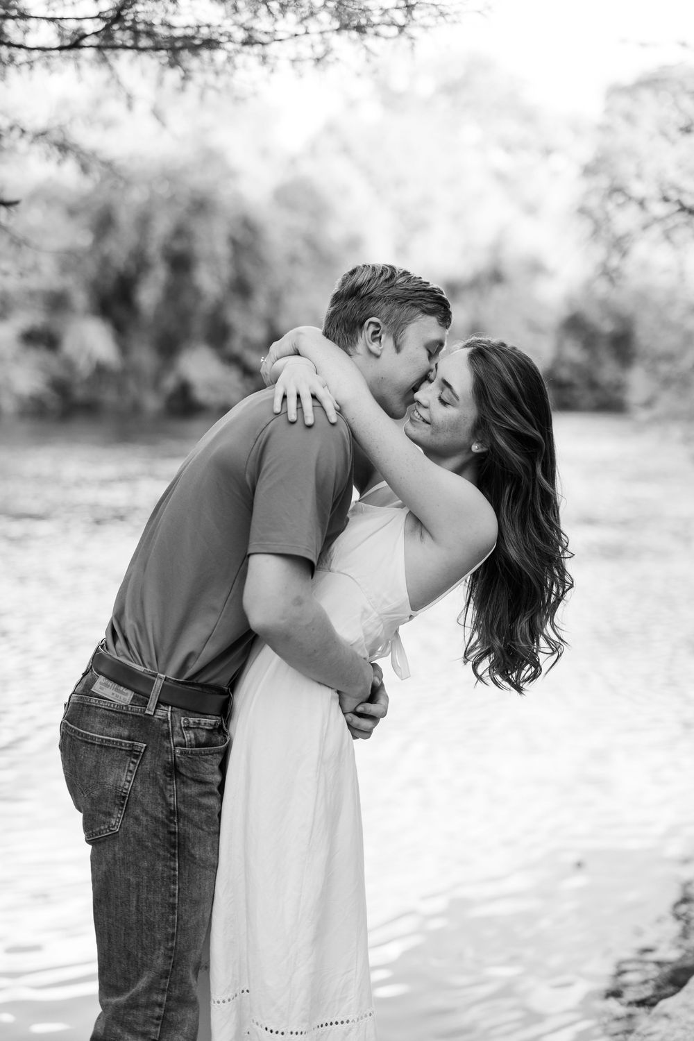 Black and white photo of couple sharing a romantic embrace near a river, woman in white dress.