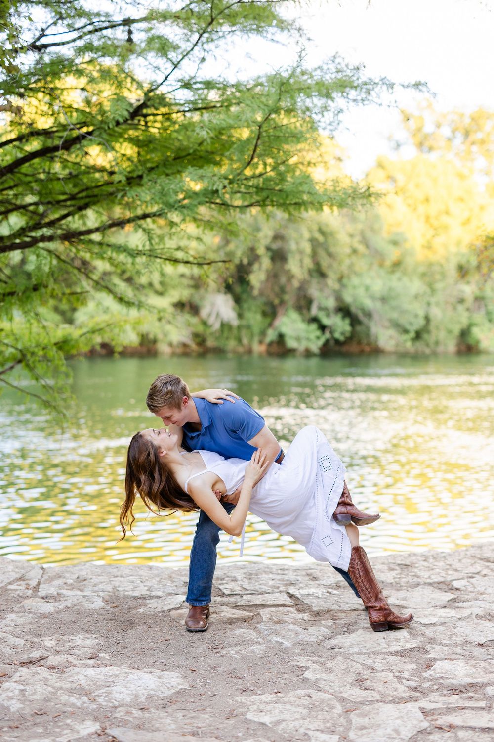 Couple sharing a romantic dip kiss by a sunlit lake, woman in white dress and cowboy boots, man in blue shirt.