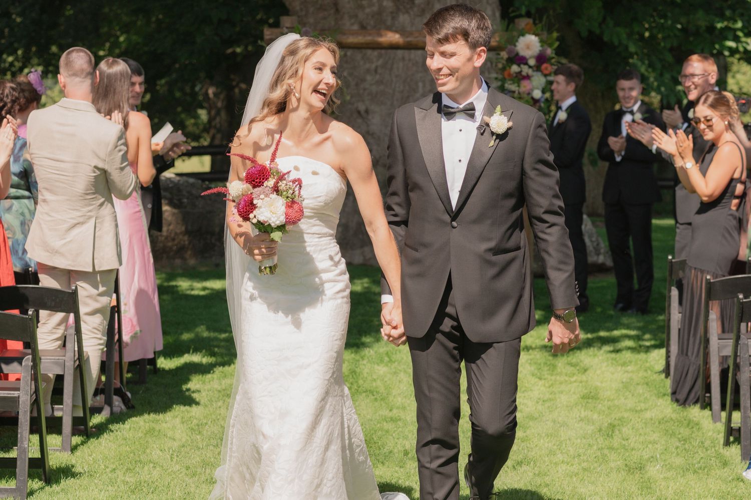 Newlywed couple walks down outdoor wedding aisle holding hands, bride in white gown with bouquet, groom in gray suit.