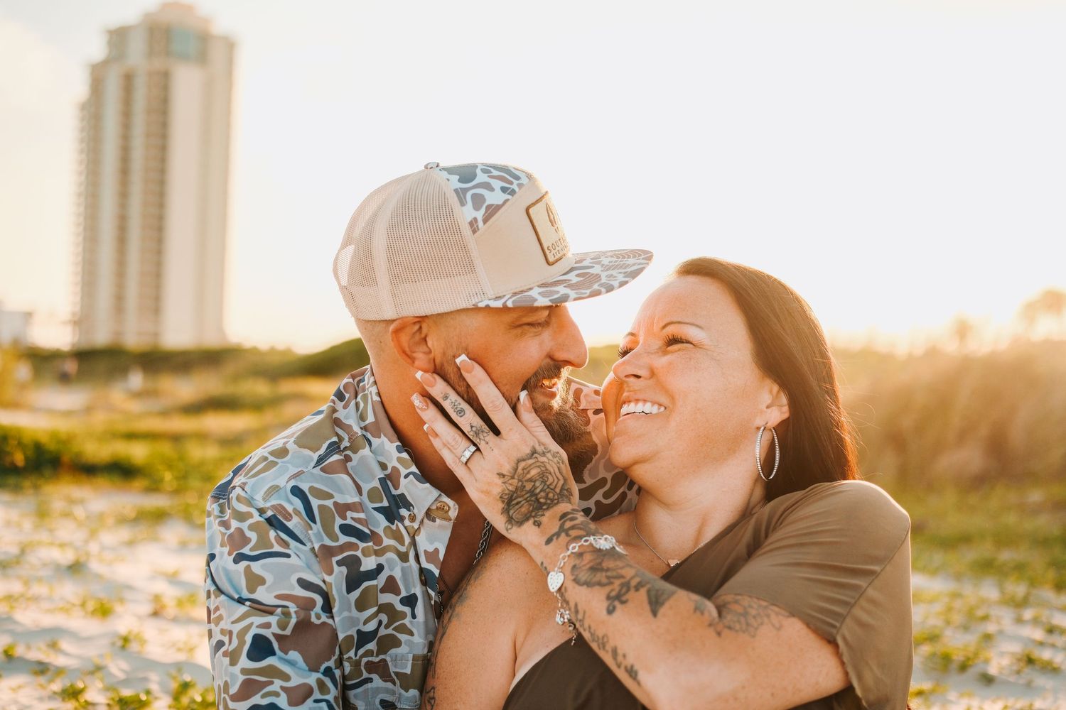 Couple sharing a romantic moment on the beach at golden hour, both with tattoos, woman smiling joyfully.