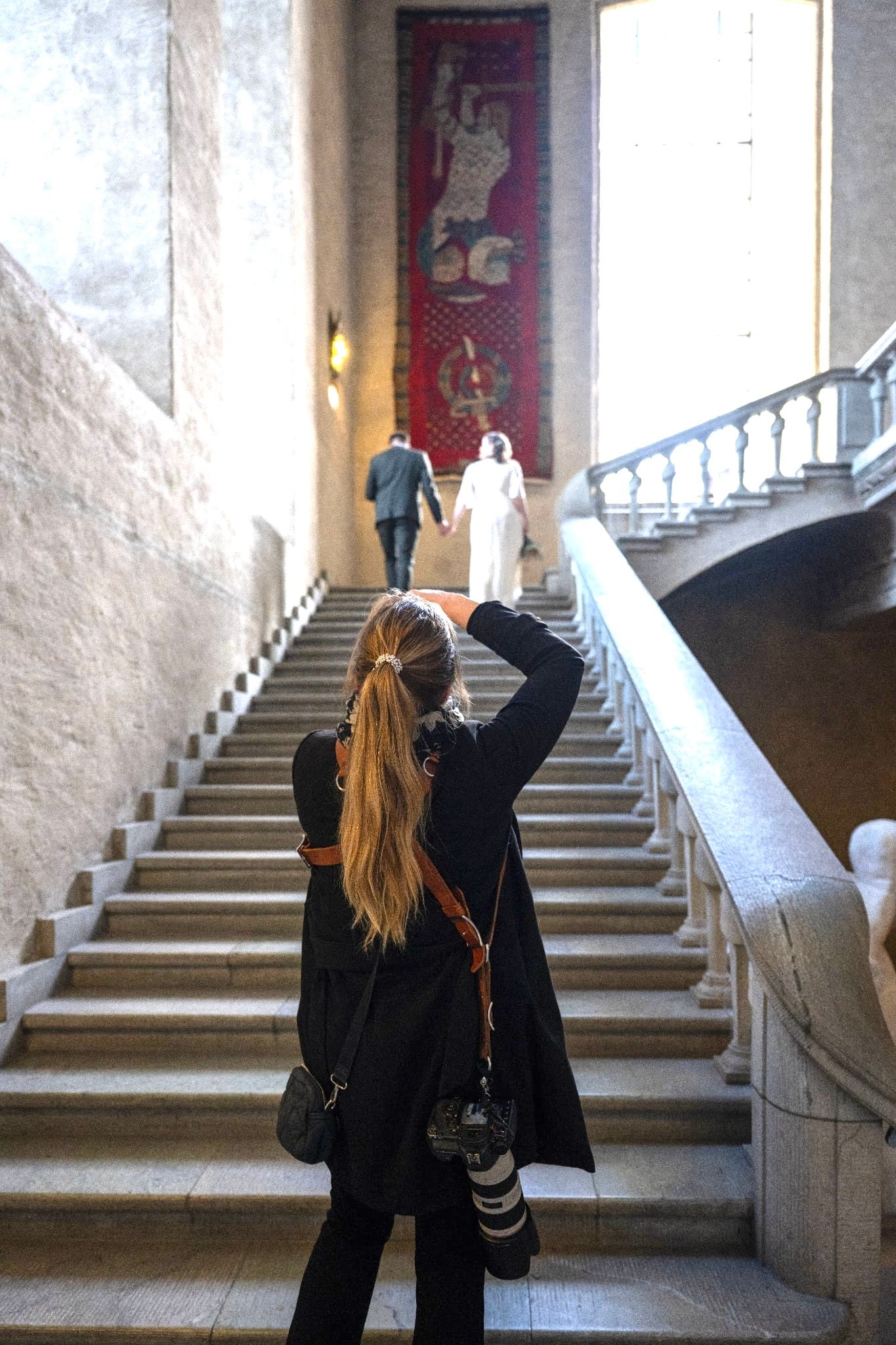 Woman photographer capturing two people ascending grand marble staircase inside ornate historic building with red tapestry.