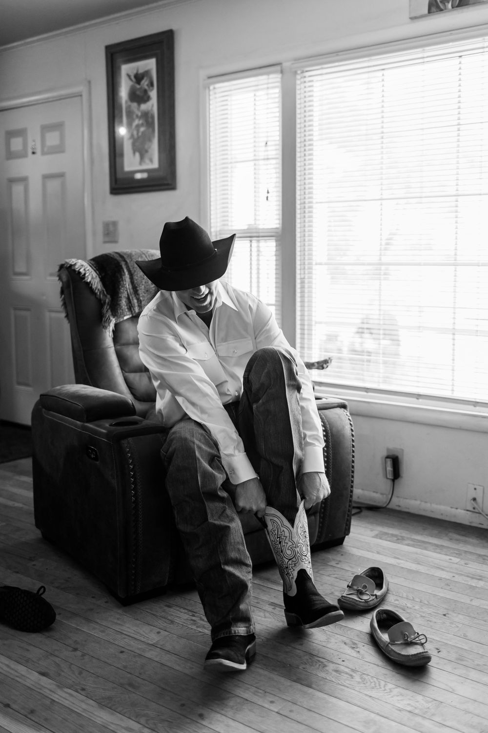 Black and white photo of a man in a cowboy hat sitting in a recliner putting on western boots in a rustic living room.